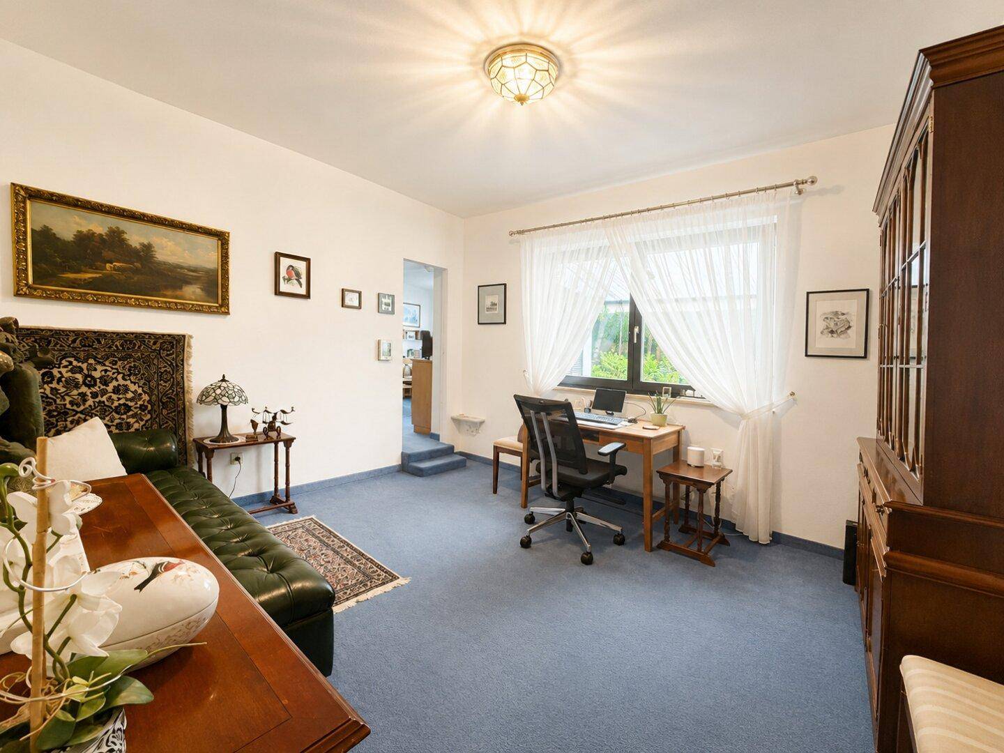 Arbeitszimmer mit Schreibtisch am Fenster, blauem Teppichboden und traditionellen Möbeln.