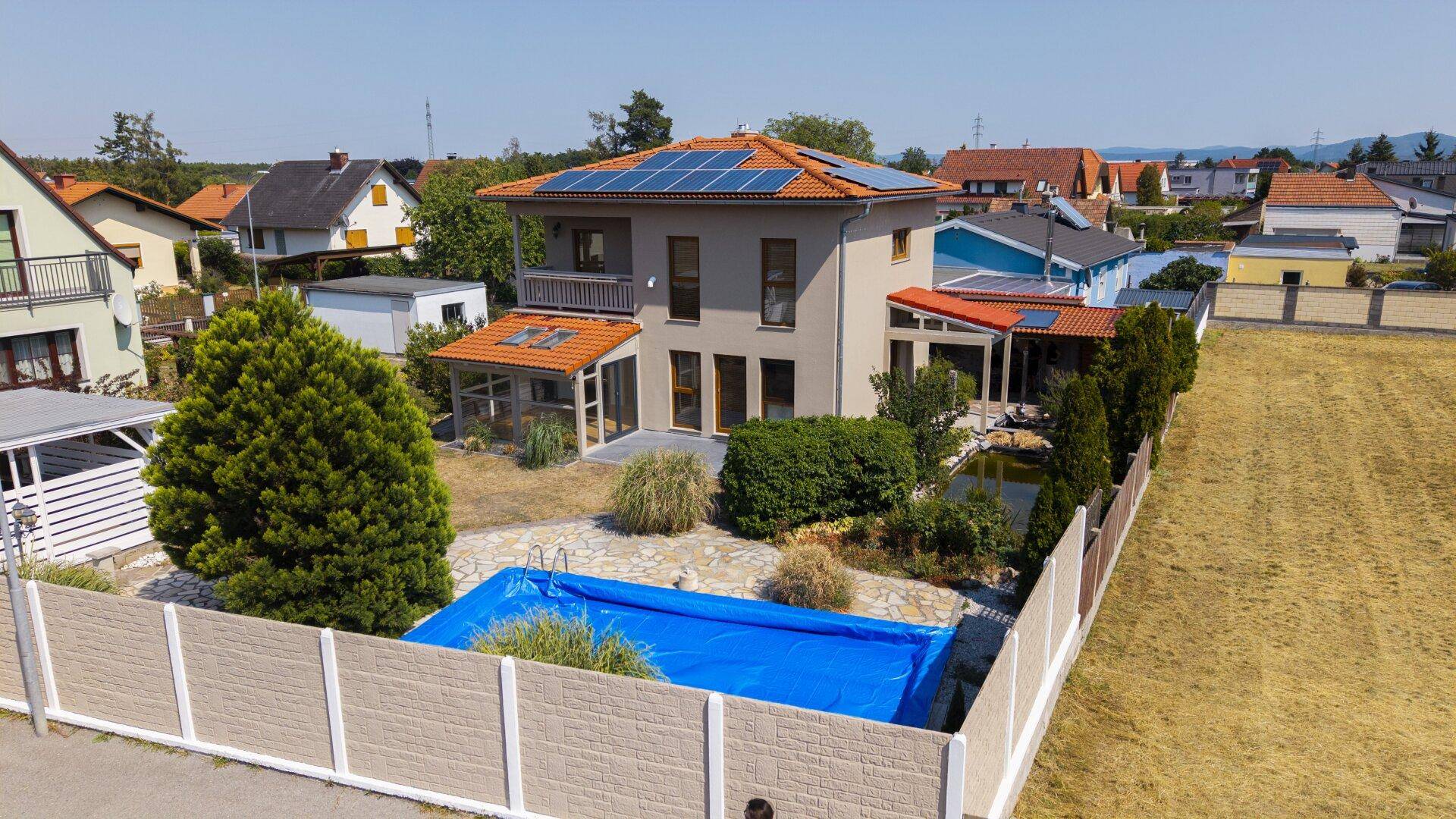 Luftaufnahme eines Einfamilienhauses mit Garten, überdachtem Pool und Photovoltaikanlage auf dem Dach.