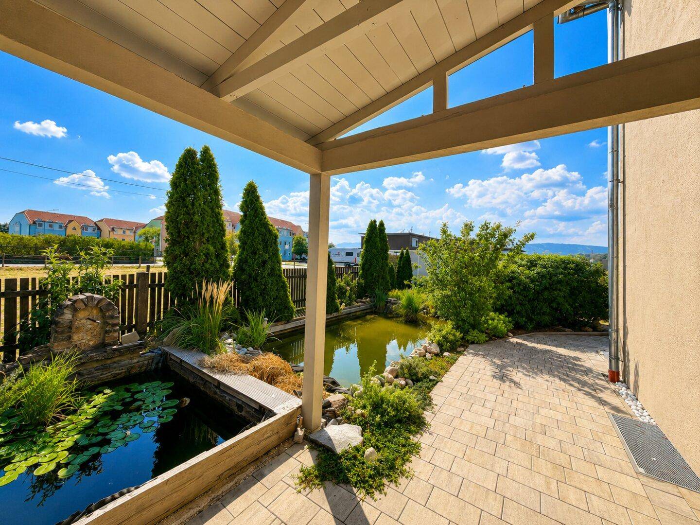 Überdachte Terrasse mit Blick auf einen Teich und den gepflegten Gartenbereich.