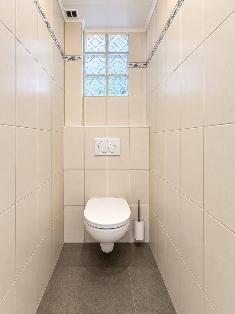 Separate Toilette mit Wand-WC und Fliesenboden, ausgestattet mit einem Glasbausteinfenster.