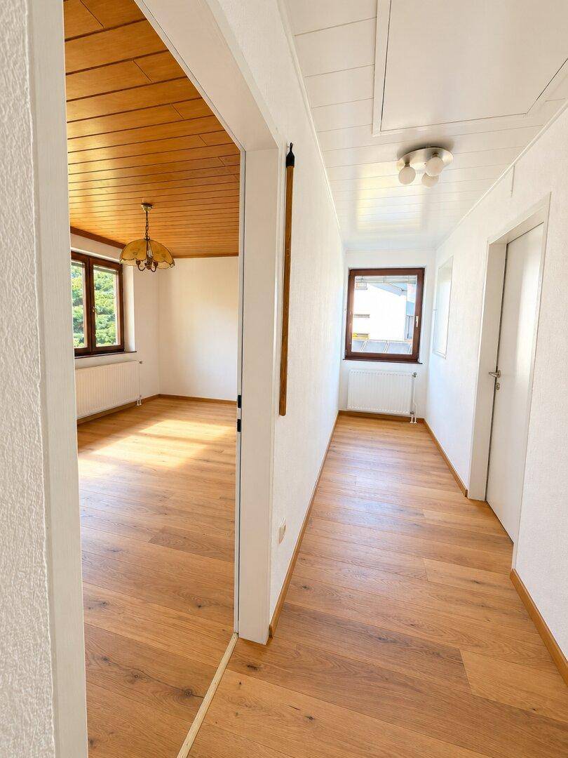 Heller Flur mit Holzboden und Blick in ein Zimmer mit Holzdecke und Fenster.