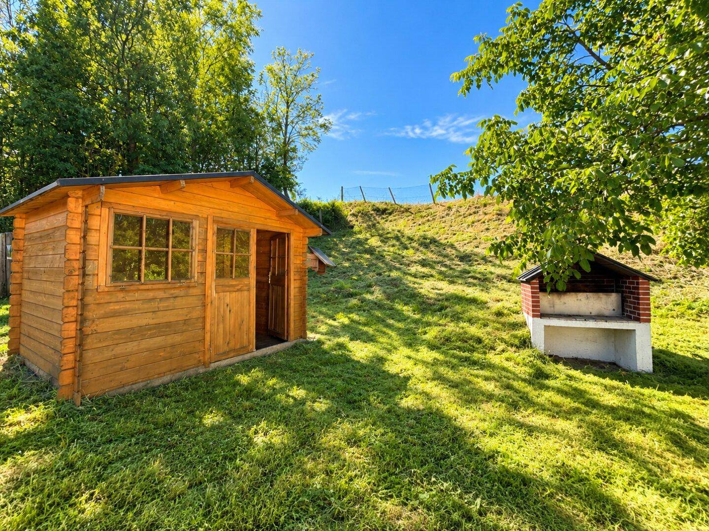 Sonniger Gartenbereich mit einem Holzschuppen und gemauertem Grillplatz im Grünen.