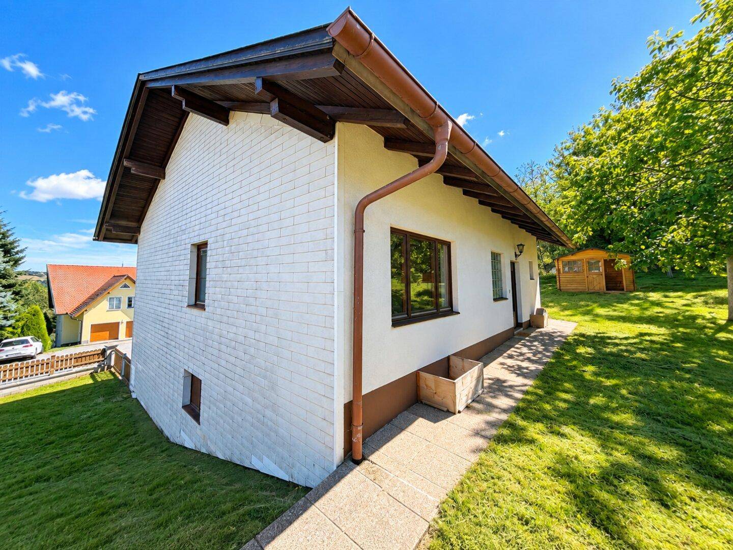 Seitenansicht des Hauses mit weißer Fassade, braunem Dach und einem gepflasterten Weg im Grünen.
