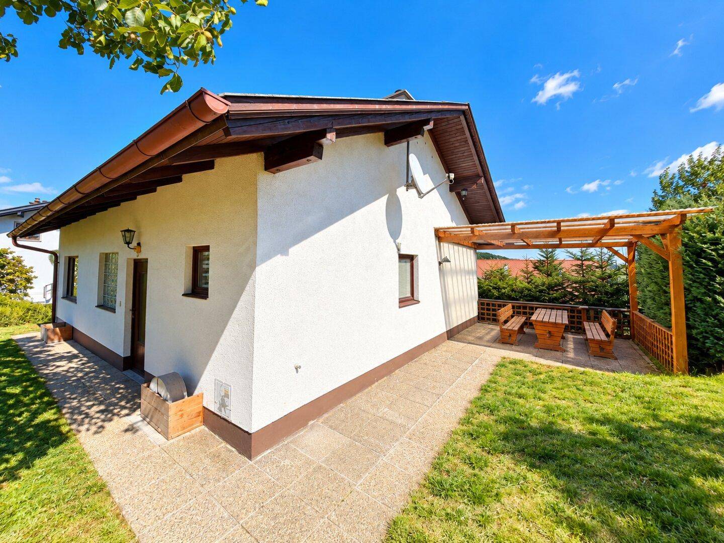 Hausfassade mit gepflastertem Weg und einer überdachten Terrasse mit Holzbänken und Tisch.