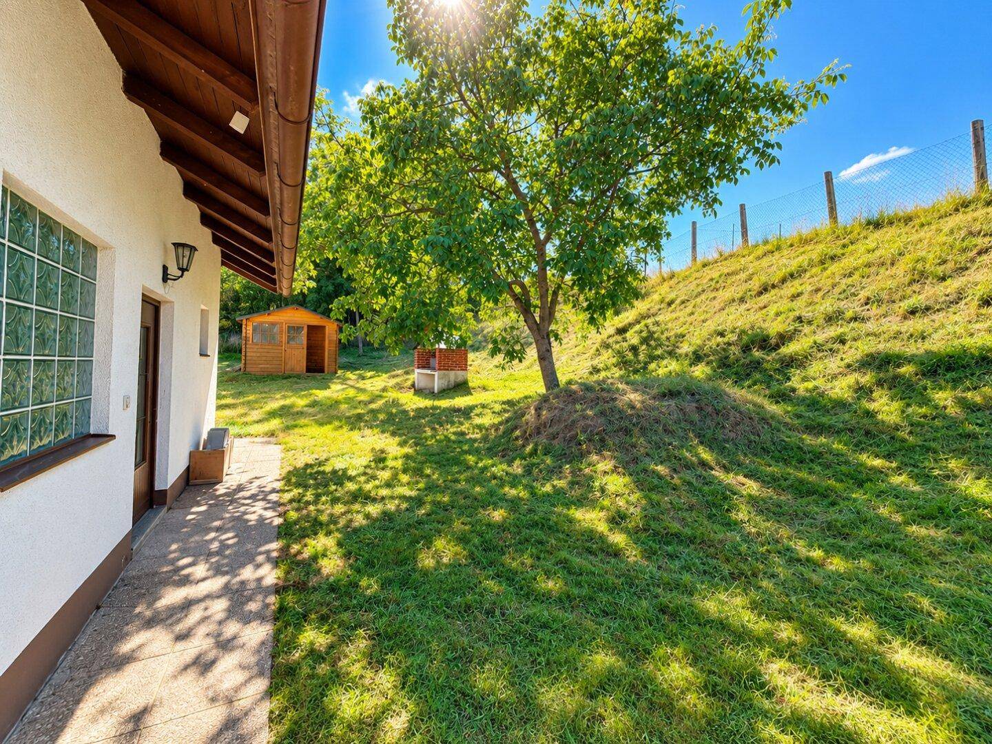 Sonniger Gartenbereich neben dem Haus mit einem Baum, Holzschuppen und Grillplatz im Hintergrund.