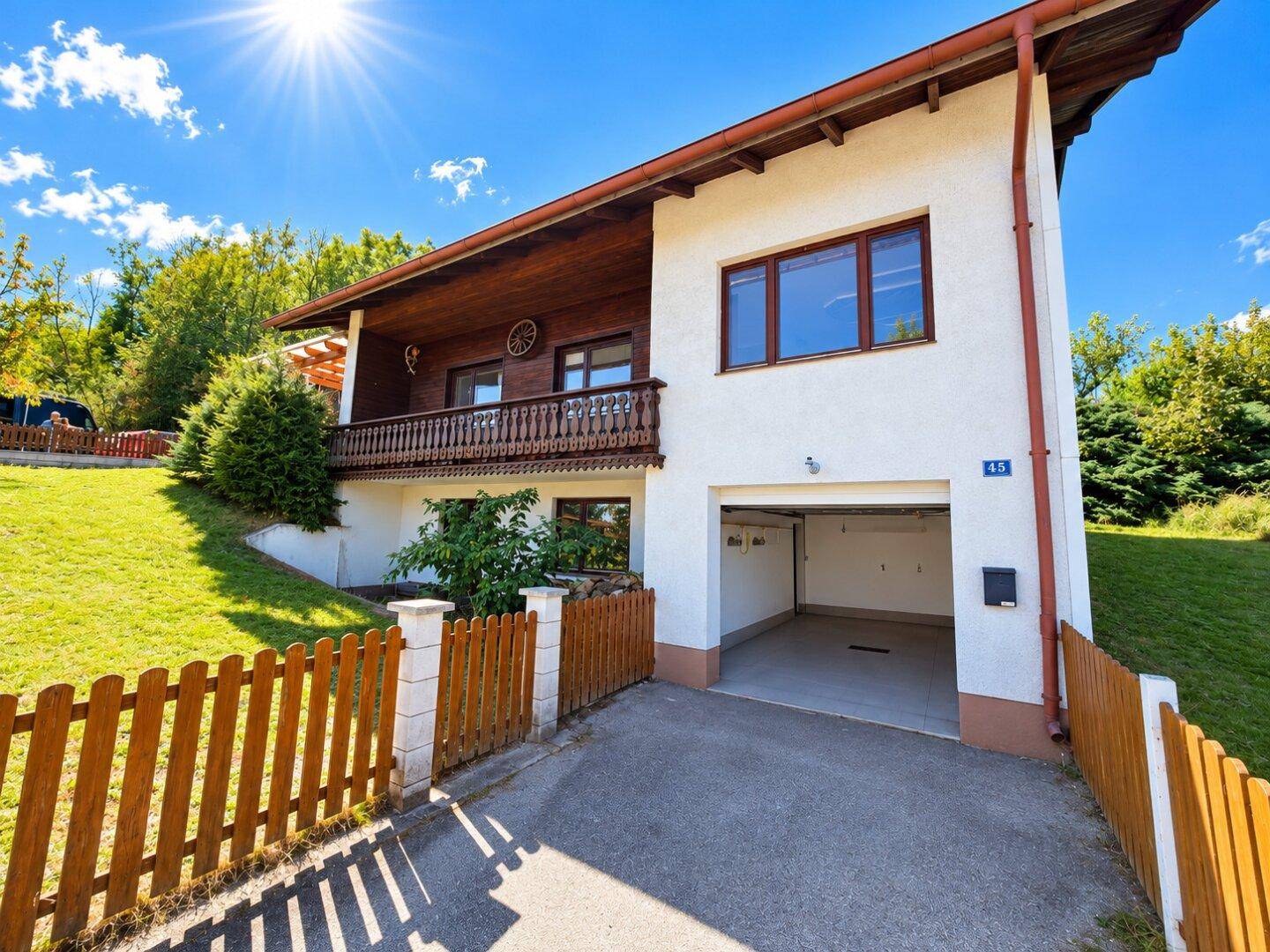 Vorderansicht des Hauses mit integrierter Garage, Balkon und Zufahrt unter strahlend blauem Himmel.