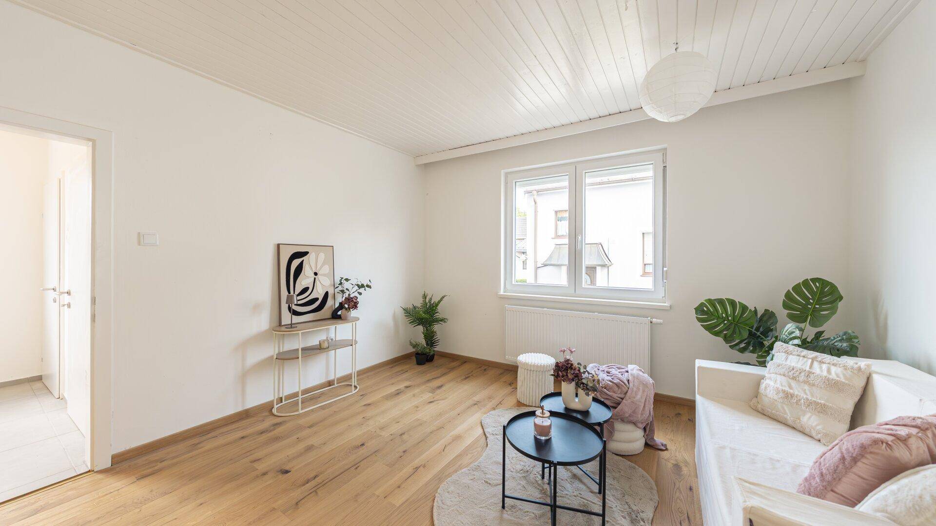 Freundliches Wohnzimmer mit Holzboden, weißer Deckenverkleidung und Blick auf die Nachbarschaft.