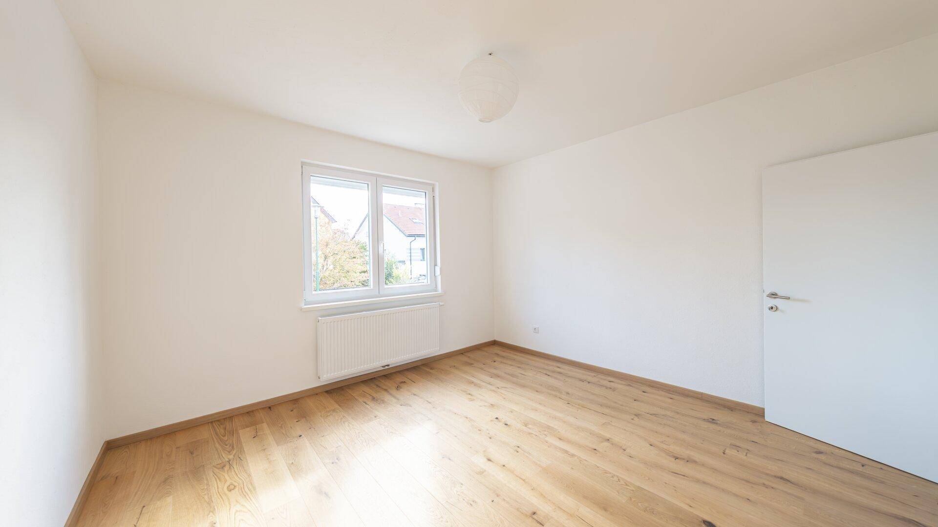 Leeres Schlafzimmer mit Holzboden, weißen Wänden und Fenster mit Blick ins Grüne.