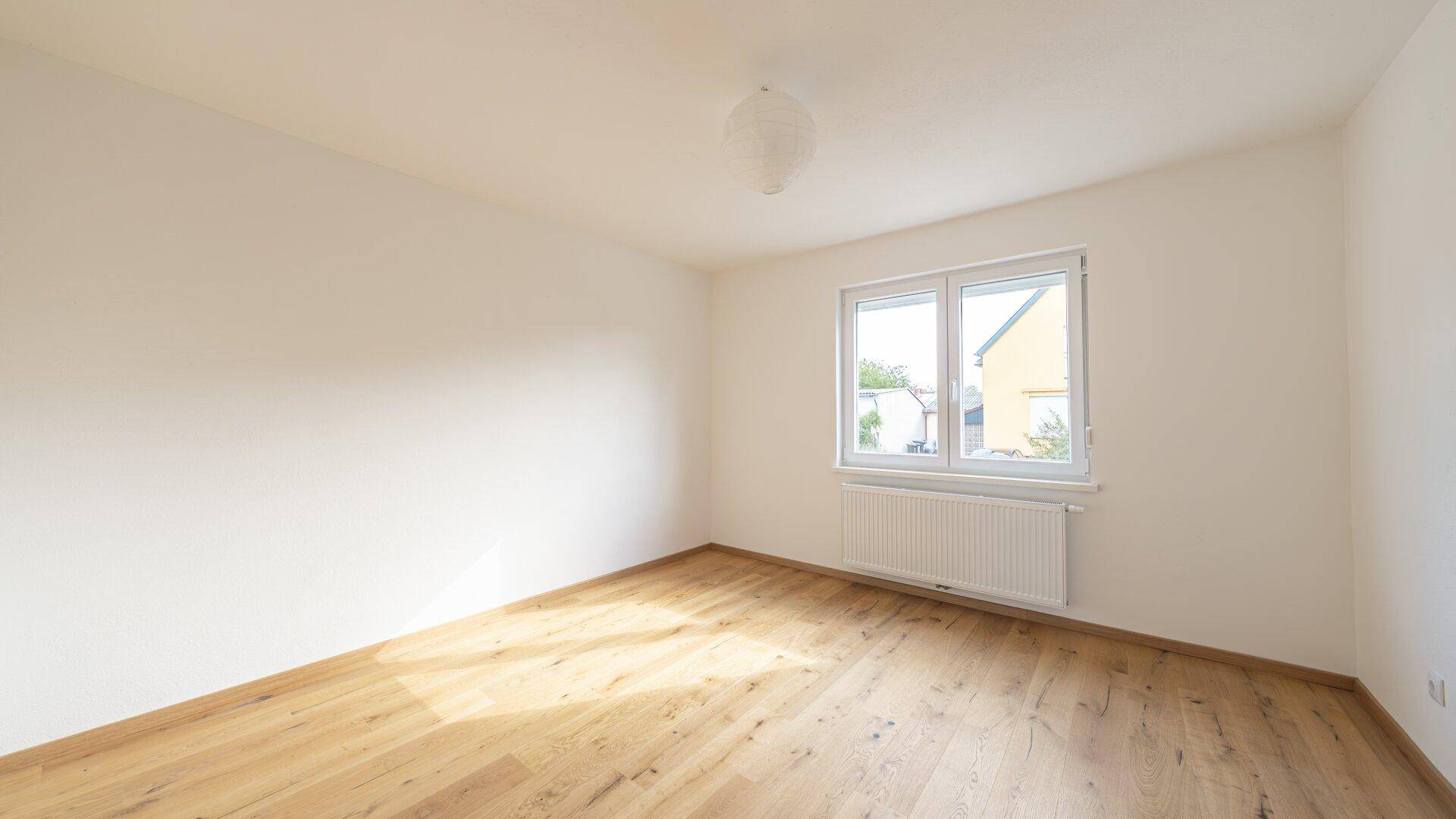 Geräumiges, leeres Zimmer mit Holzboden und weißer Wandfarbe, ideal für ein Schlafzimmer.