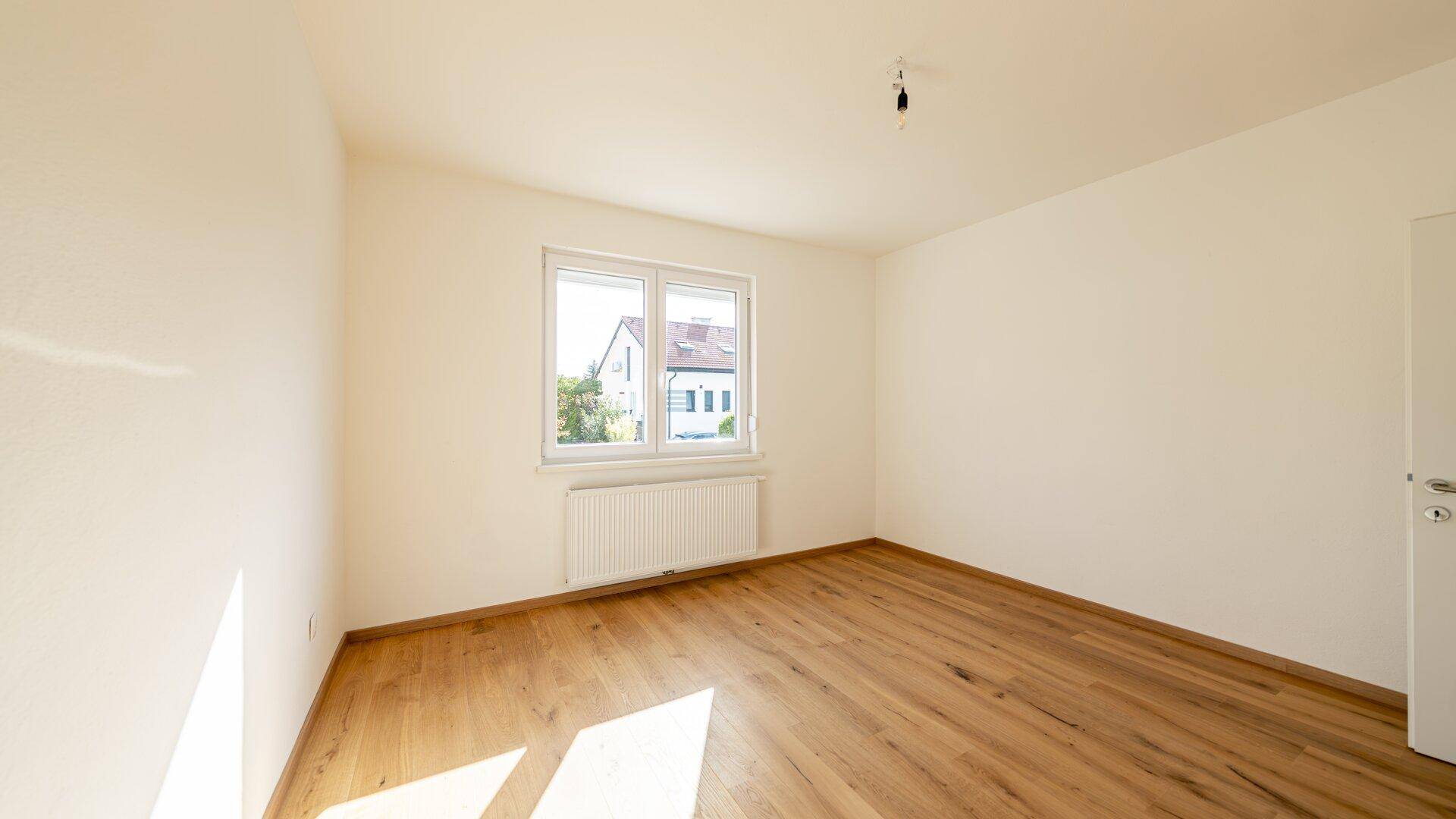 Leeres Schlafzimmer mit Holzboden, weißen Wänden und Fenster mit Blick auf die Umgebung.