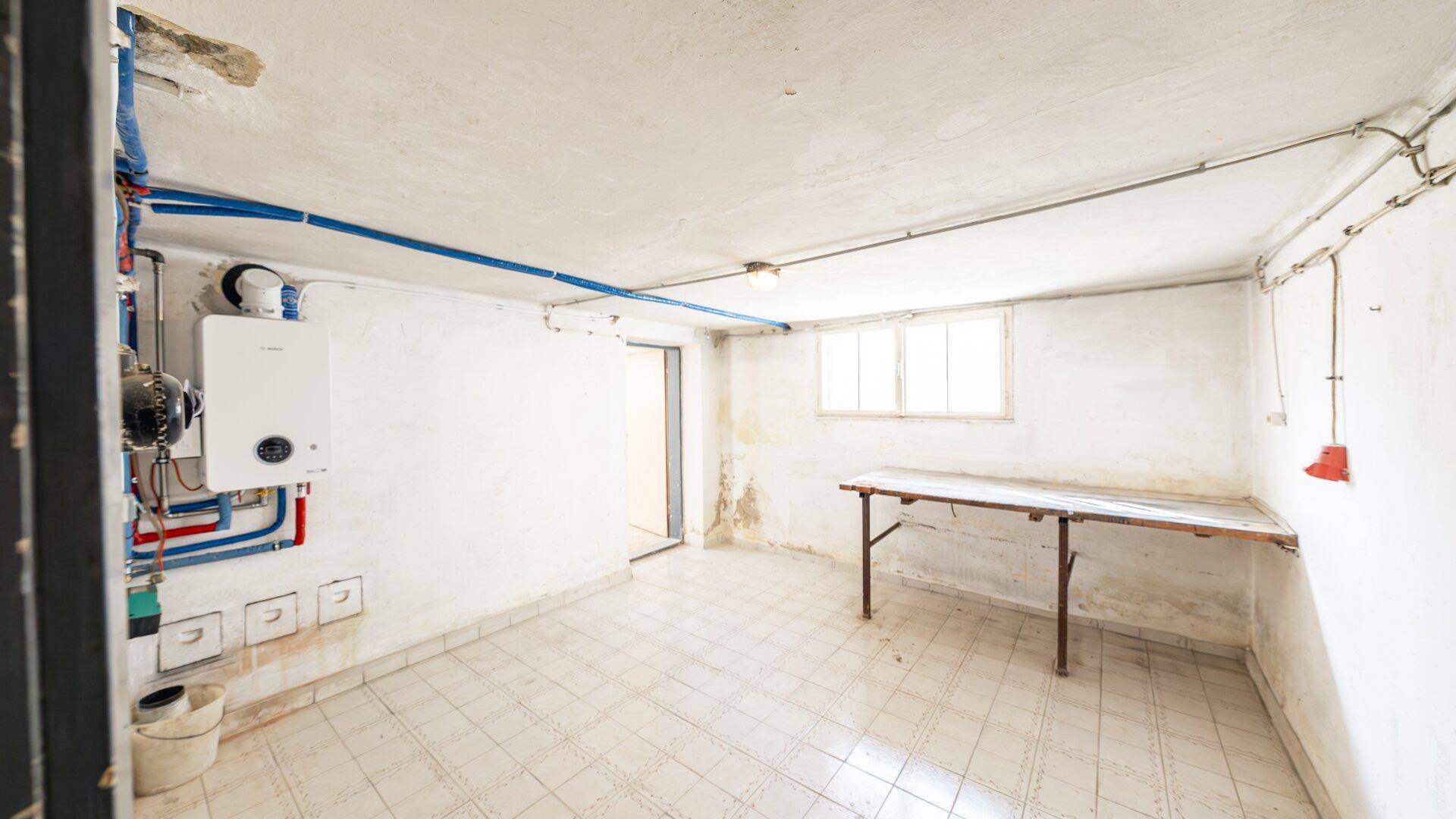 Hauswirtschaftsraum im Keller mit Warmwasserbereiter, sichtbaren Leitungen und einem Fenster, bietet Arbeitsfläche.