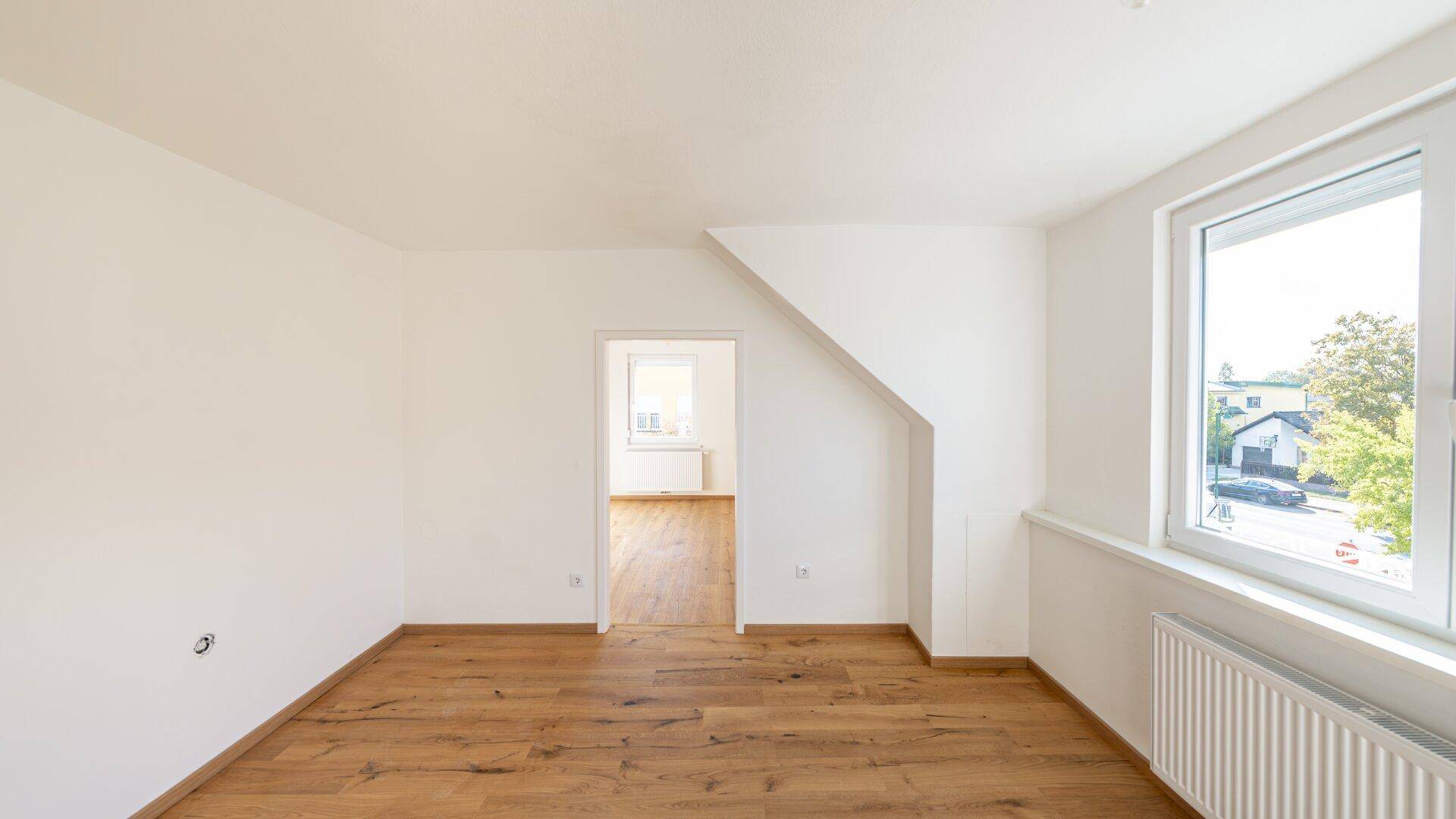 Helles Zimmer im Dachgeschoss mit Holzboden, Schrägdach und Blick auf die Umgebung.