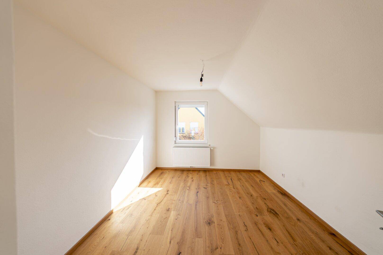 Leeres Zimmer im Dachgeschoss mit Holzboden, Schrägdach und Fenster.
