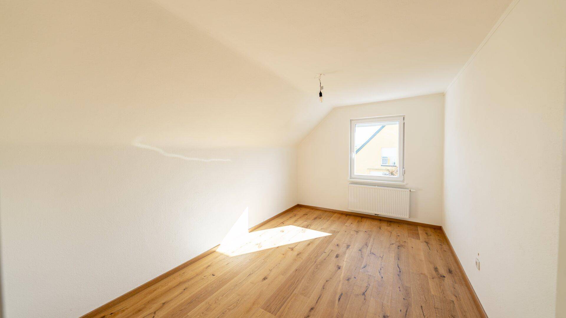 Helles Zimmer im Dachgeschoss mit Holzboden, Schrägdach und Fenster.