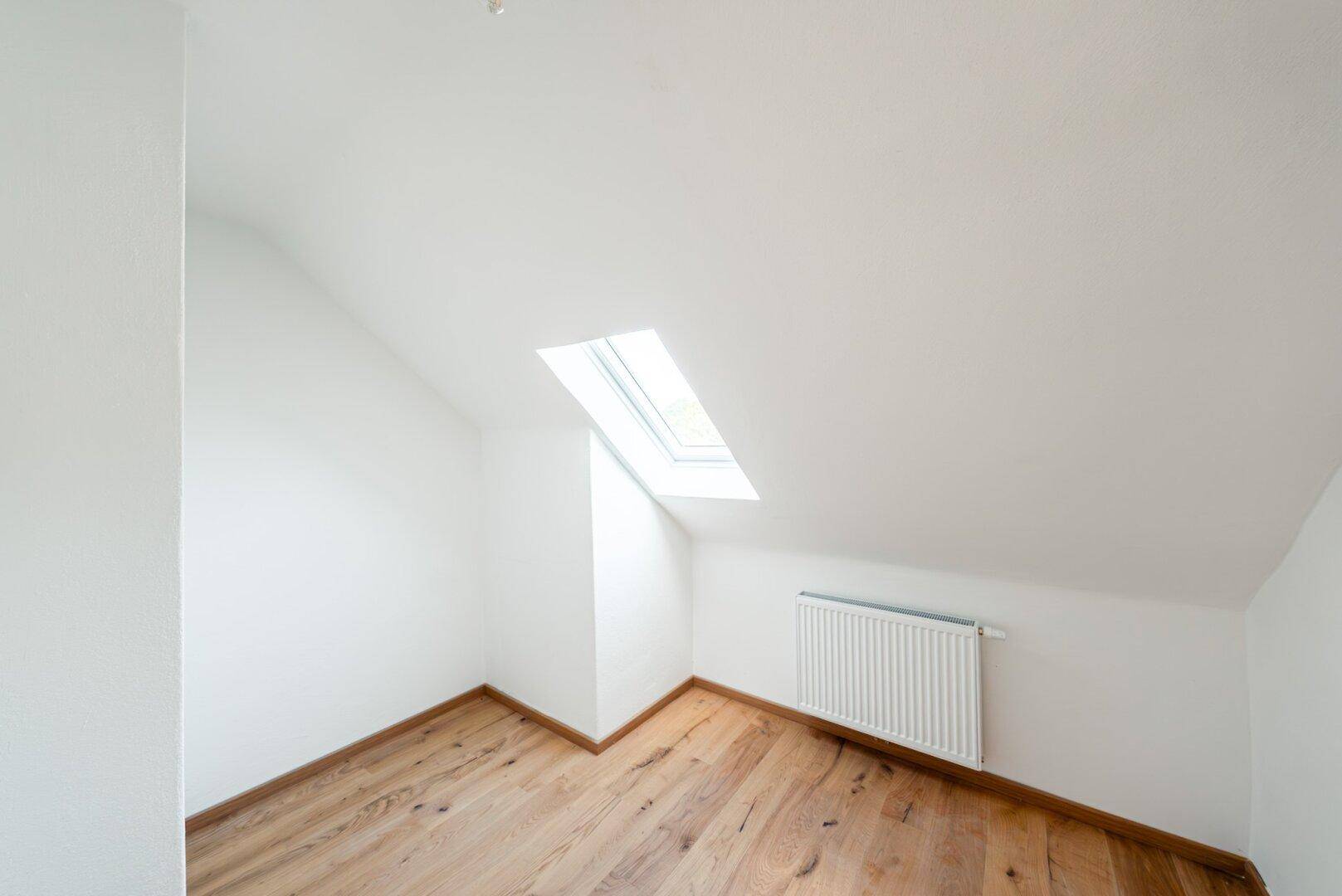 Dachgeschosszimmer mit Holzboden, weiß gestrichenen Wänden und Dachfenster für Tageslicht.