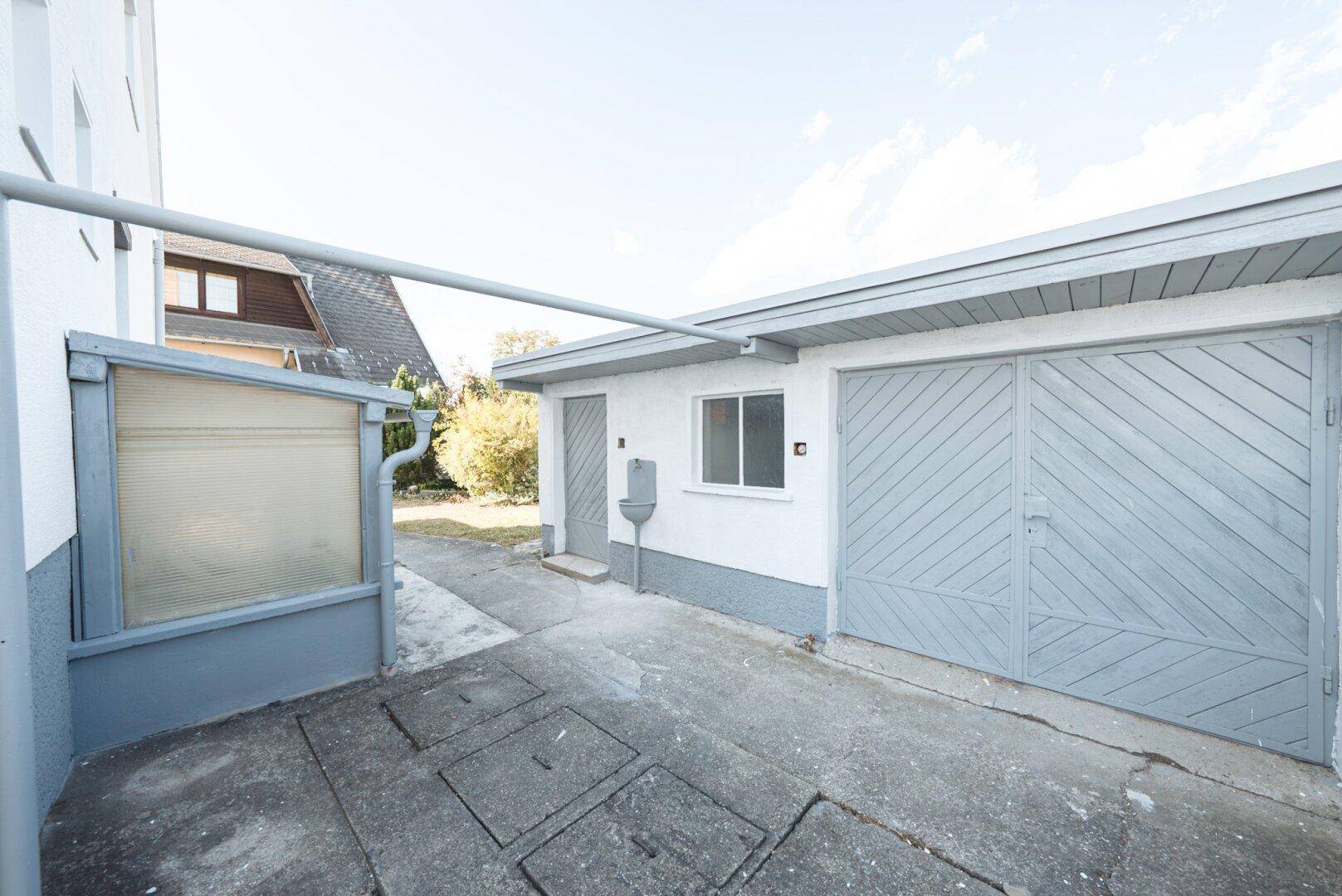 Garage mit Doppeltor, Seiteneingang und einem kleinen überdachten Bereich, umgeben von gepflastertem Boden.