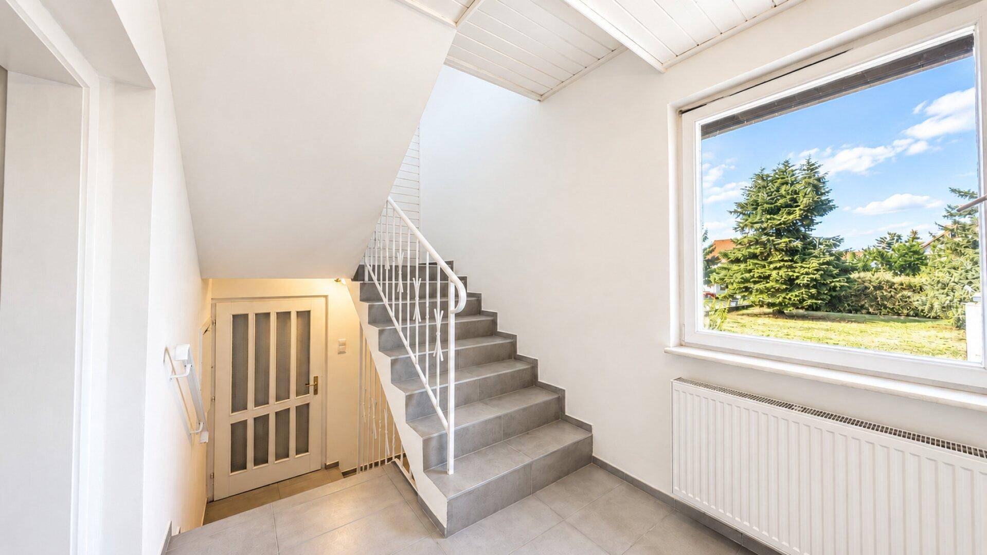 Heller Treppenaufgang mit Fliesenboden, weißem Geländer und Fenster mit Blick ins Grüne.