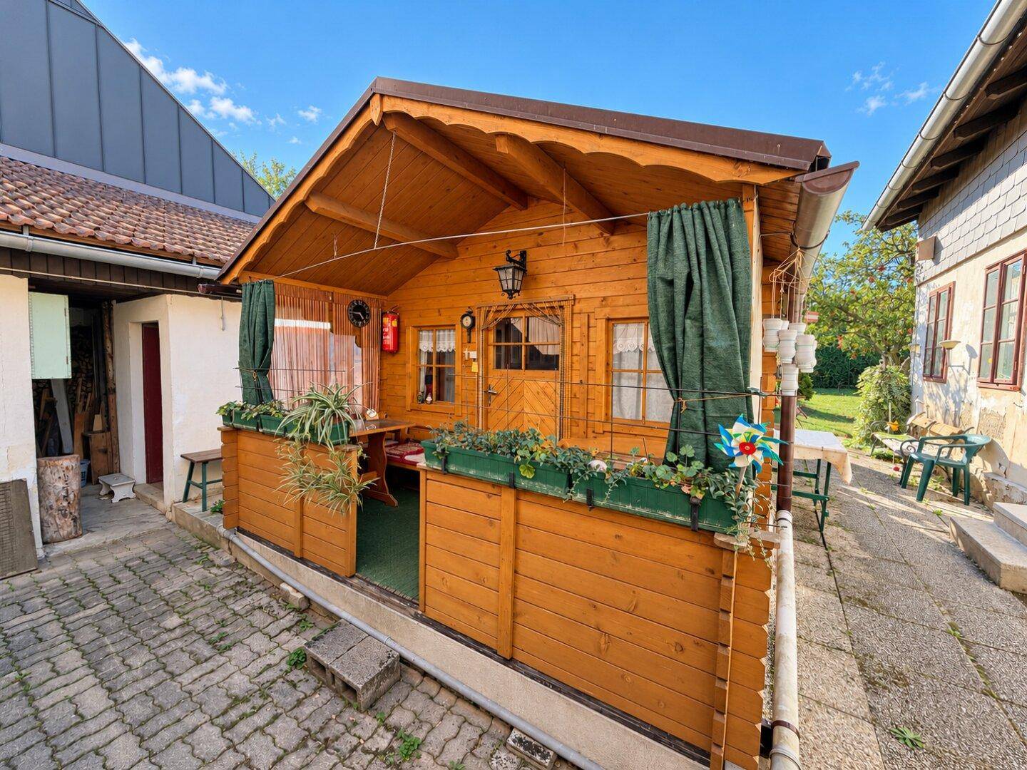 Kleine Holzhütte mit überdachter Terrasse, bepflanzten Blumenkästen und Sitzgelegenheiten im Freien.