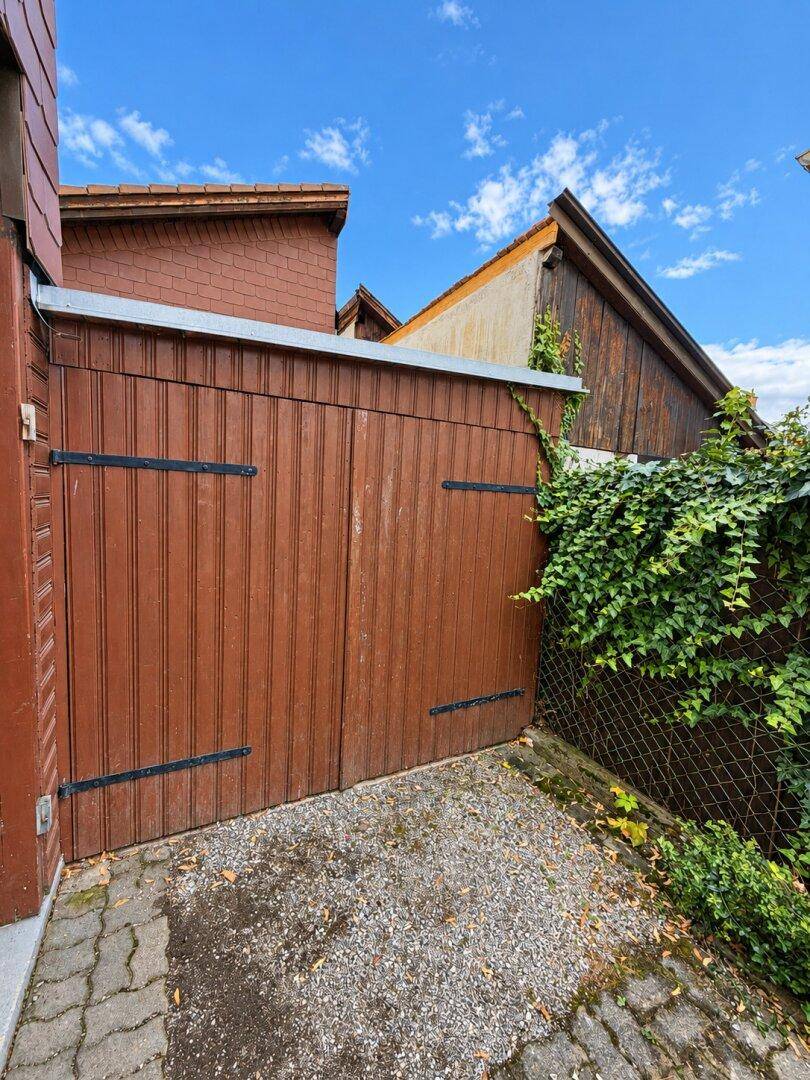 Braunes Holztor einer Garage, umgeben von Efeu und einer gepflasterten Einfahrt.