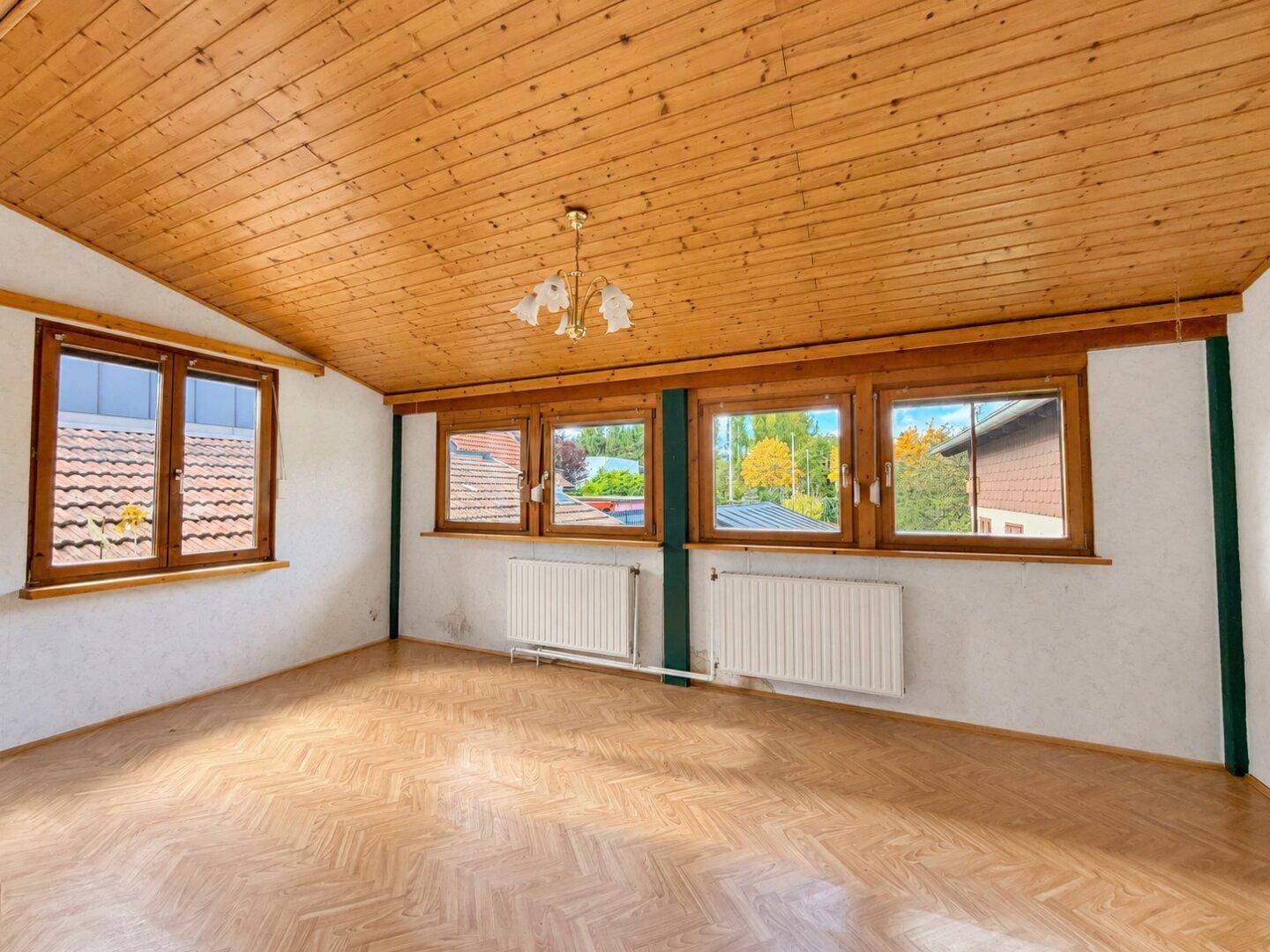 Geräumiges Zimmer mit Holzdecke, Laminatboden und mehreren Fenstern, die viel Licht hereinlassen.