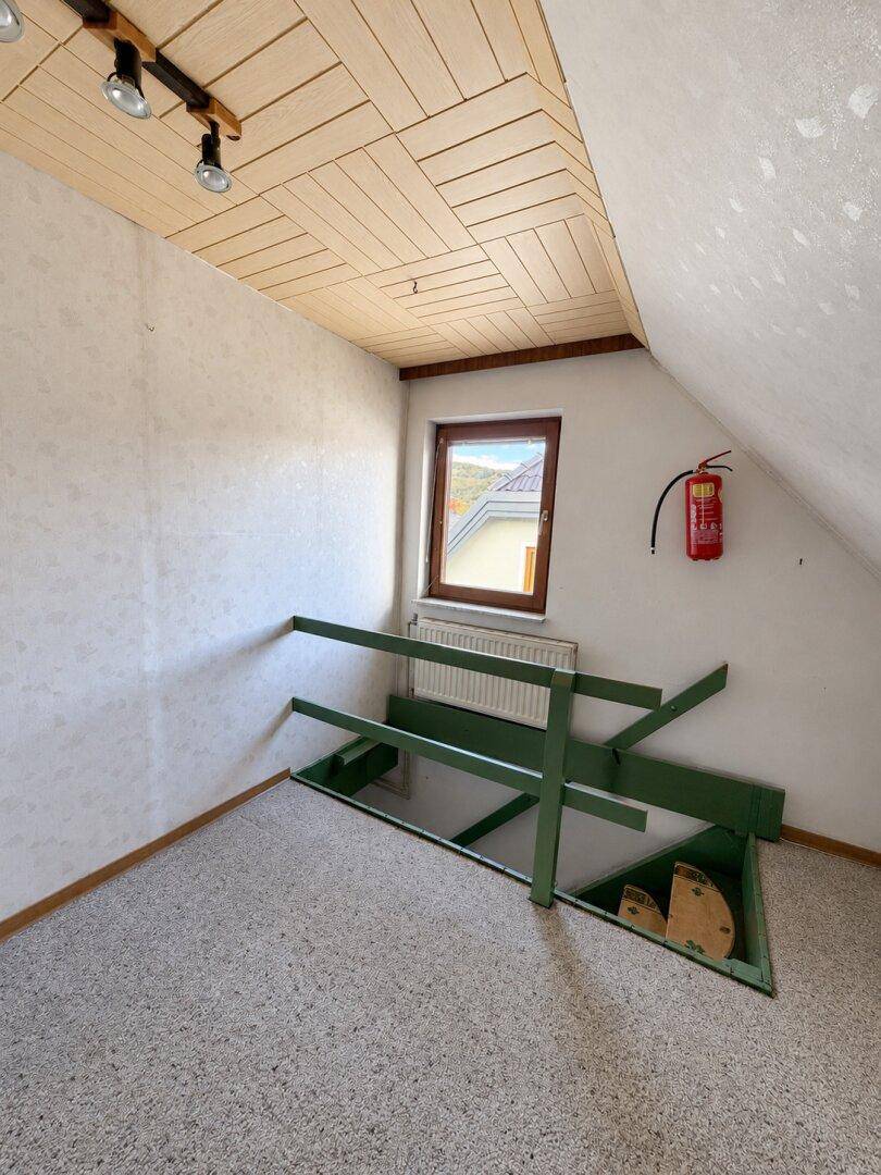 Dachgeschossraum mit Teppichboden, schrägen Wänden und einem kleinen Fenster.