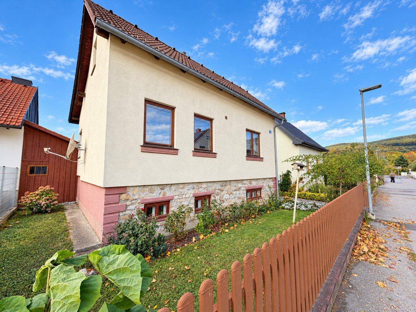Seitenansicht des Hauses mit Vorgarten, Holzzaun und Blick auf die ländliche Umgebung und Straße.