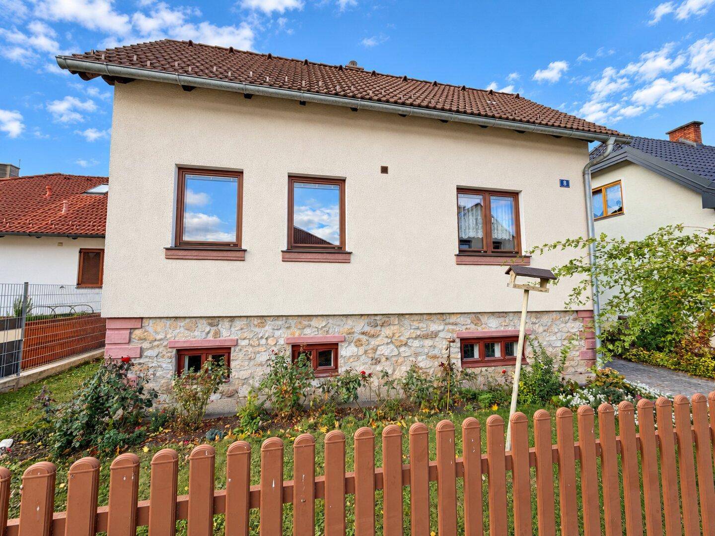 Vorderansicht des Hauses mit beiger Fassade, braunen Fenstern und einem kleinen Vorgarten mit Holzzaun.