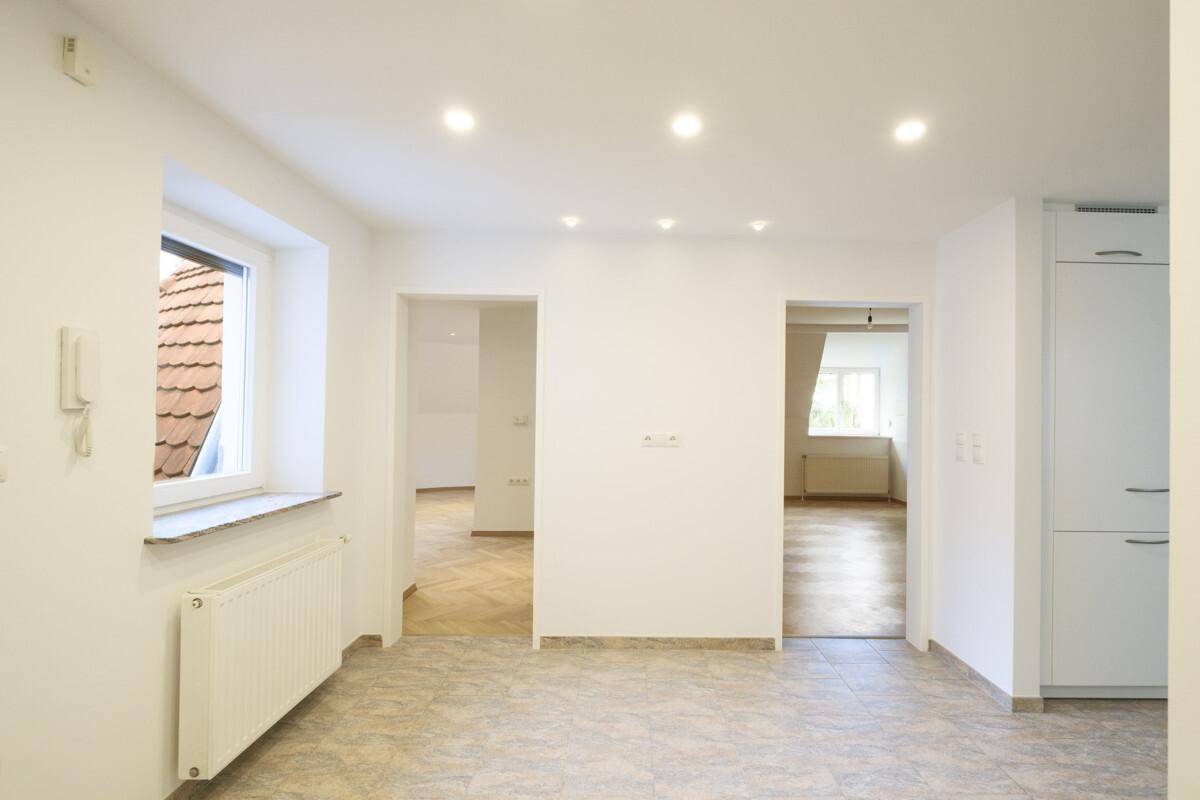 Heller Raum mit Fliesenboden, Fensterblick auf ein Ziegeldach und integrierter Deckenbeleuchtung.