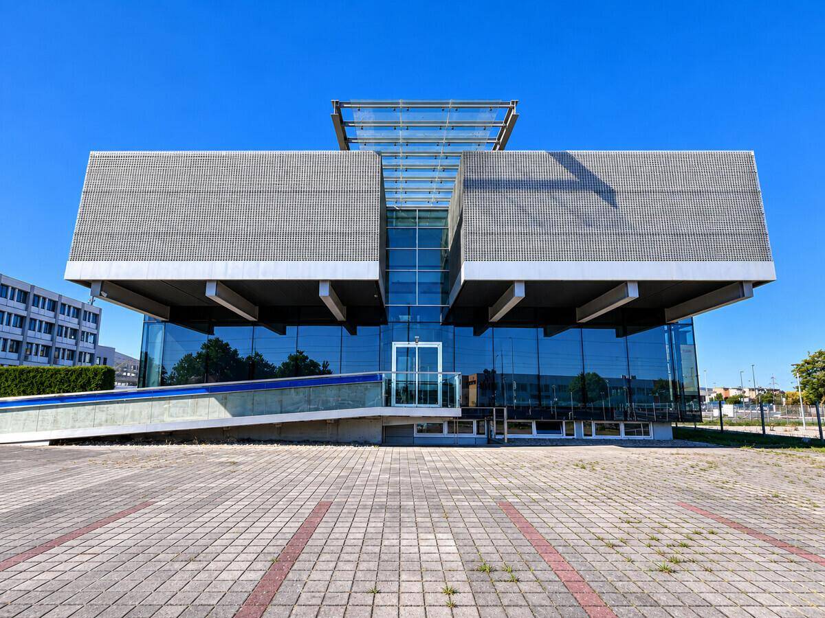 Frontansicht eines modernen Bürogebäudes mit großer Glasfront und Rampe an einem sonnigen Tag.