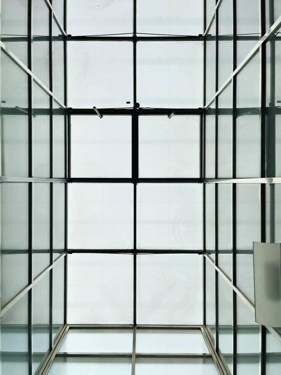 Blick nach oben auf eine moderne Glasdachkonstruktion mit Metallrahmen, die viel Licht hereinlässt.
