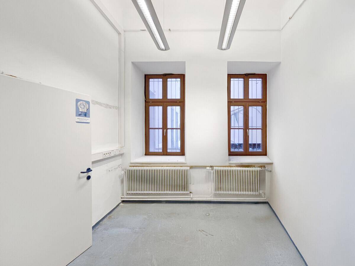 Kleinerer, heller Gewerberaum mit zwei Fenstern, Holzrahmen, Sicherheitsgittern und Heizkörpern unter den Fenstern.
