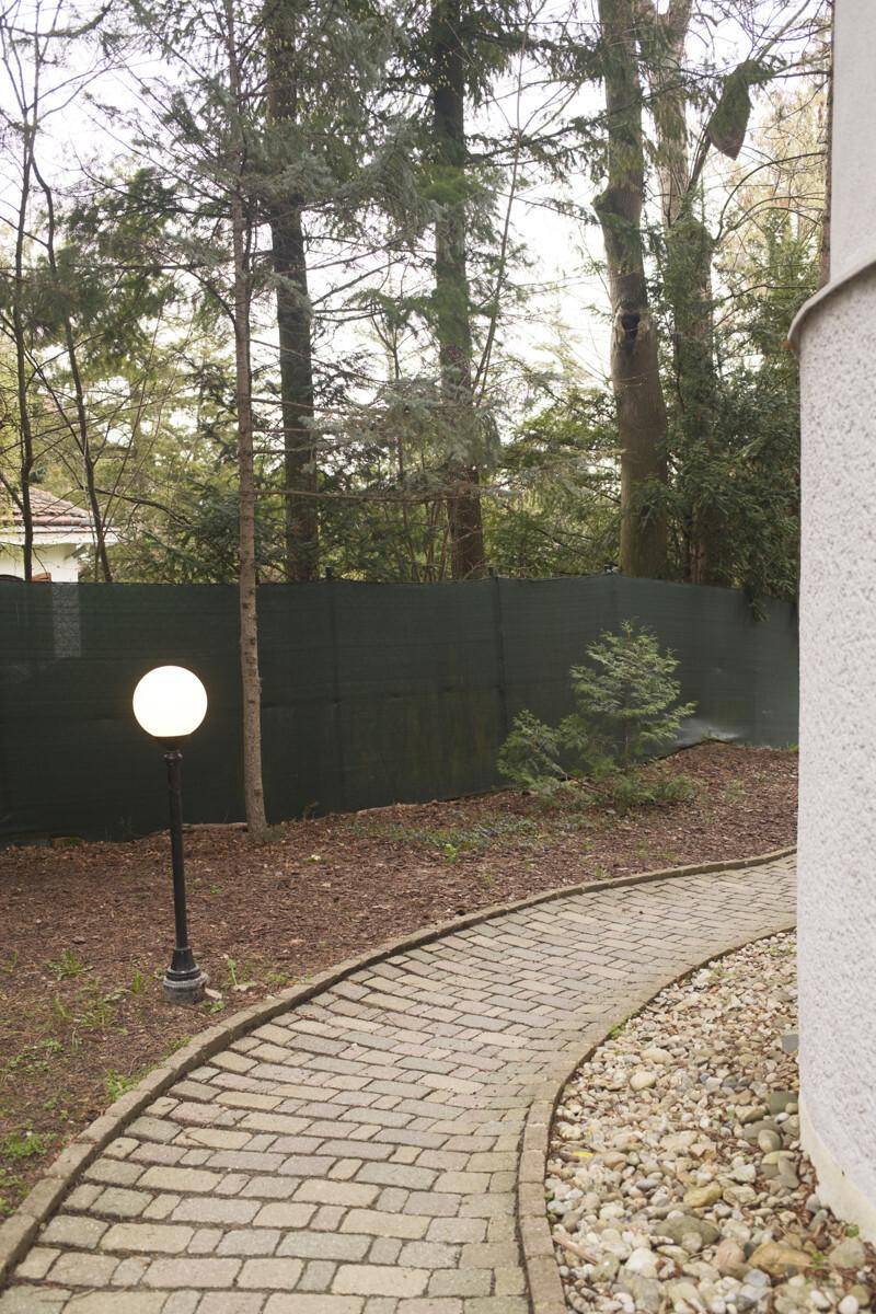 Geschwungener Pflasterweg führt durch einen Gartenbereich mit Bäumen und einer Gartenleuchte.