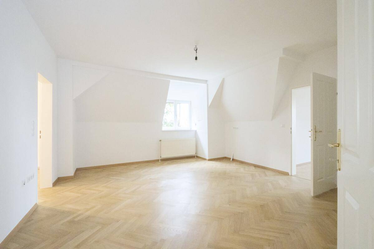 Helles Zimmer mit Fischgrätparkettboden, weißen Wänden, Fenster und Heizkörper unter der Dachschräge.