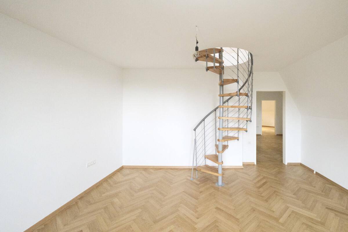 Heller Raum mit Fischgrätparkettboden, weißen Wänden und einer eleganten Wendeltreppe.
