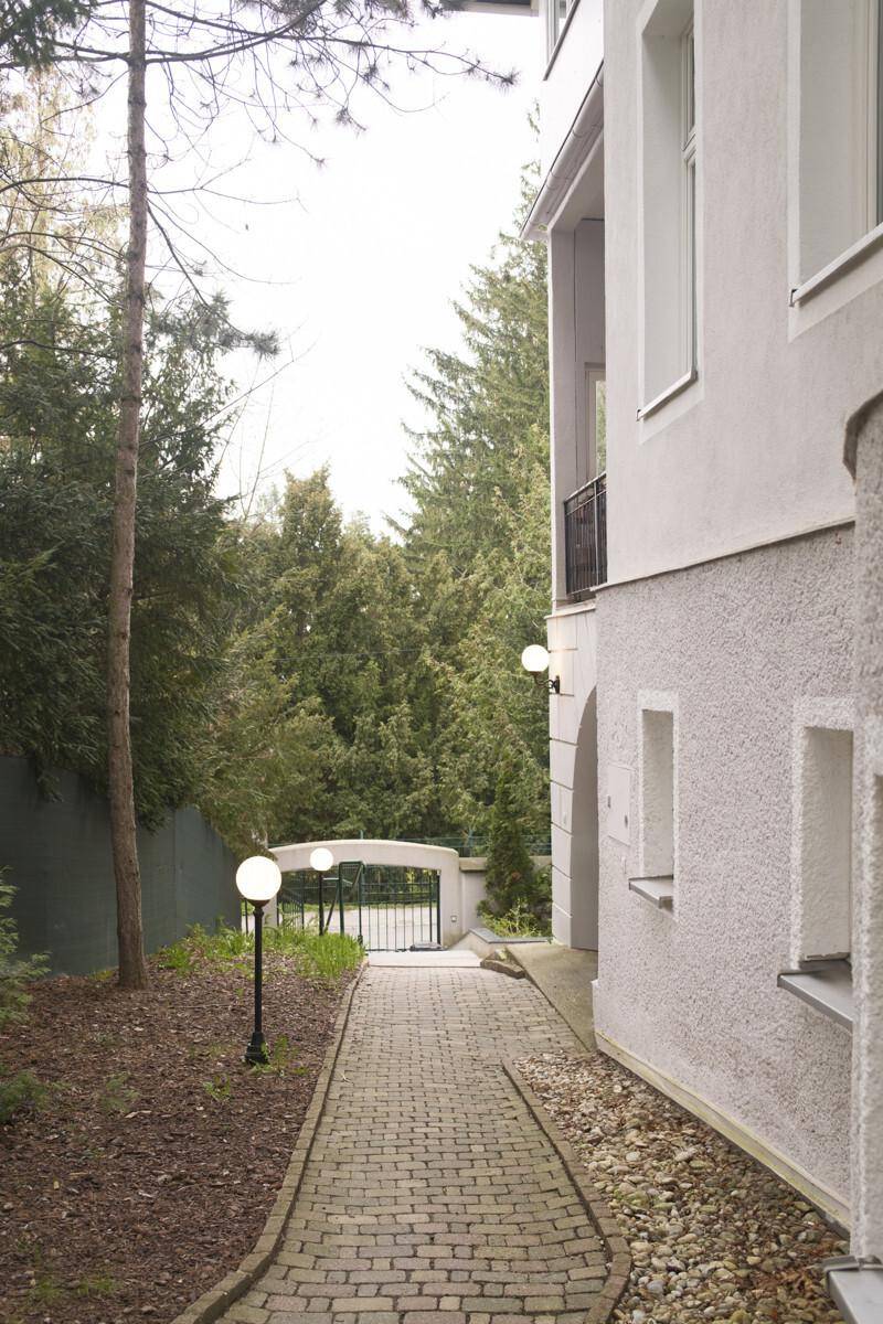 Pflasterweg führt zu einem Tor, umgeben von Bäumen und einer Gebäudefassade mit Außenleuchten.