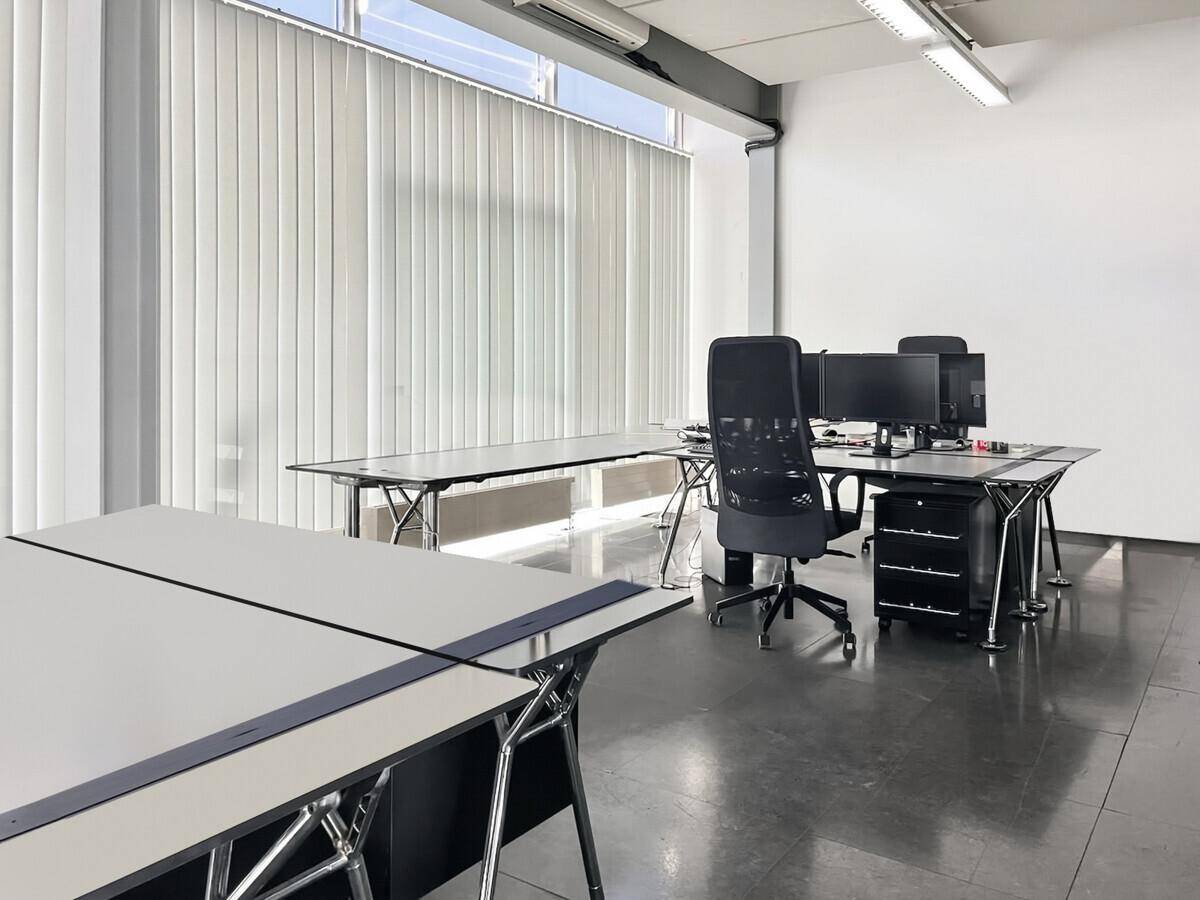 Geräumiges Büro mit mehreren Schreibtischen, vertikalen Jalousien an großen Fenstern und dunklem Boden.
