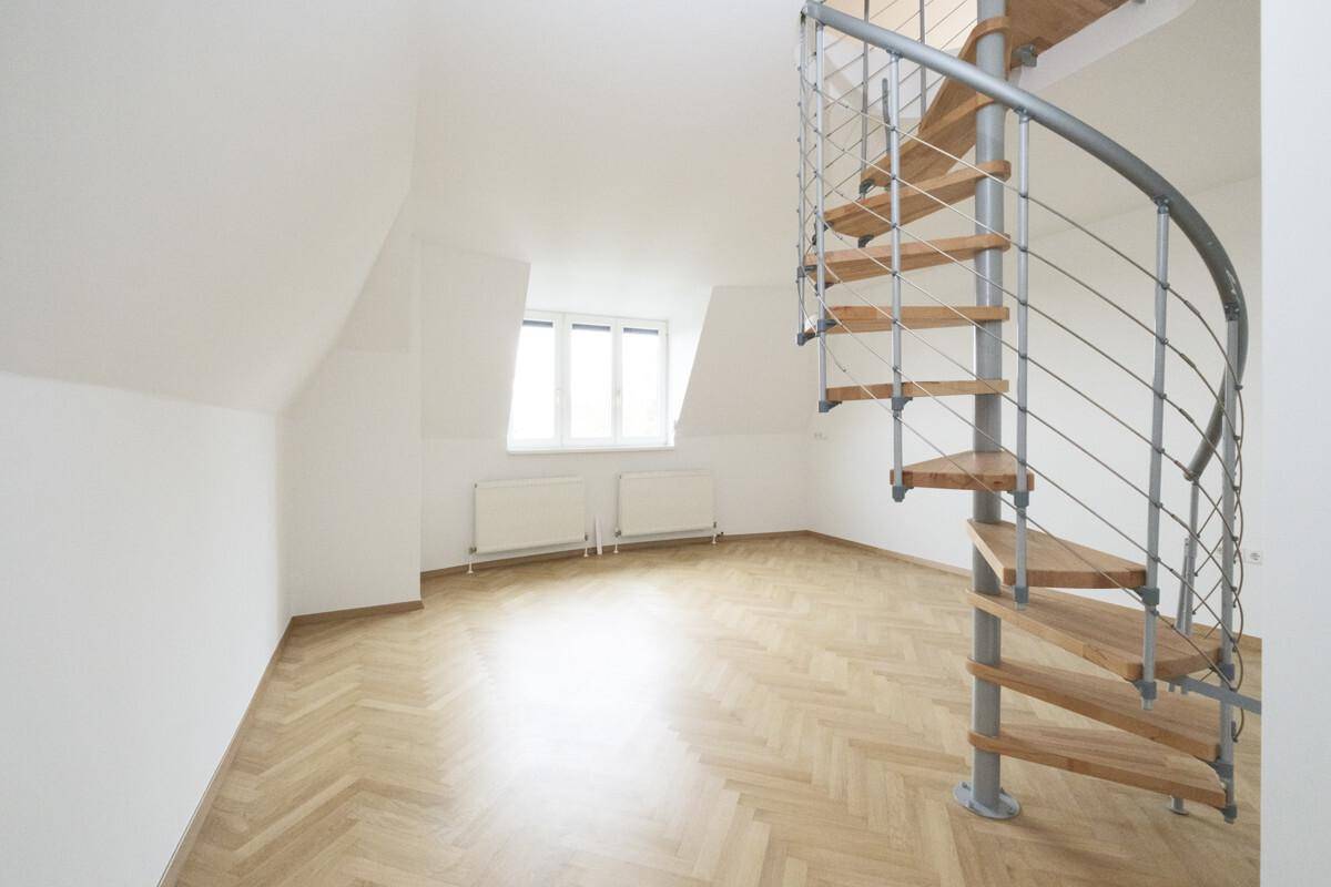 Geräumiger Raum mit Fischgrätparkett, Fenster, Heizkörpern und einer markanten Wendeltreppe.