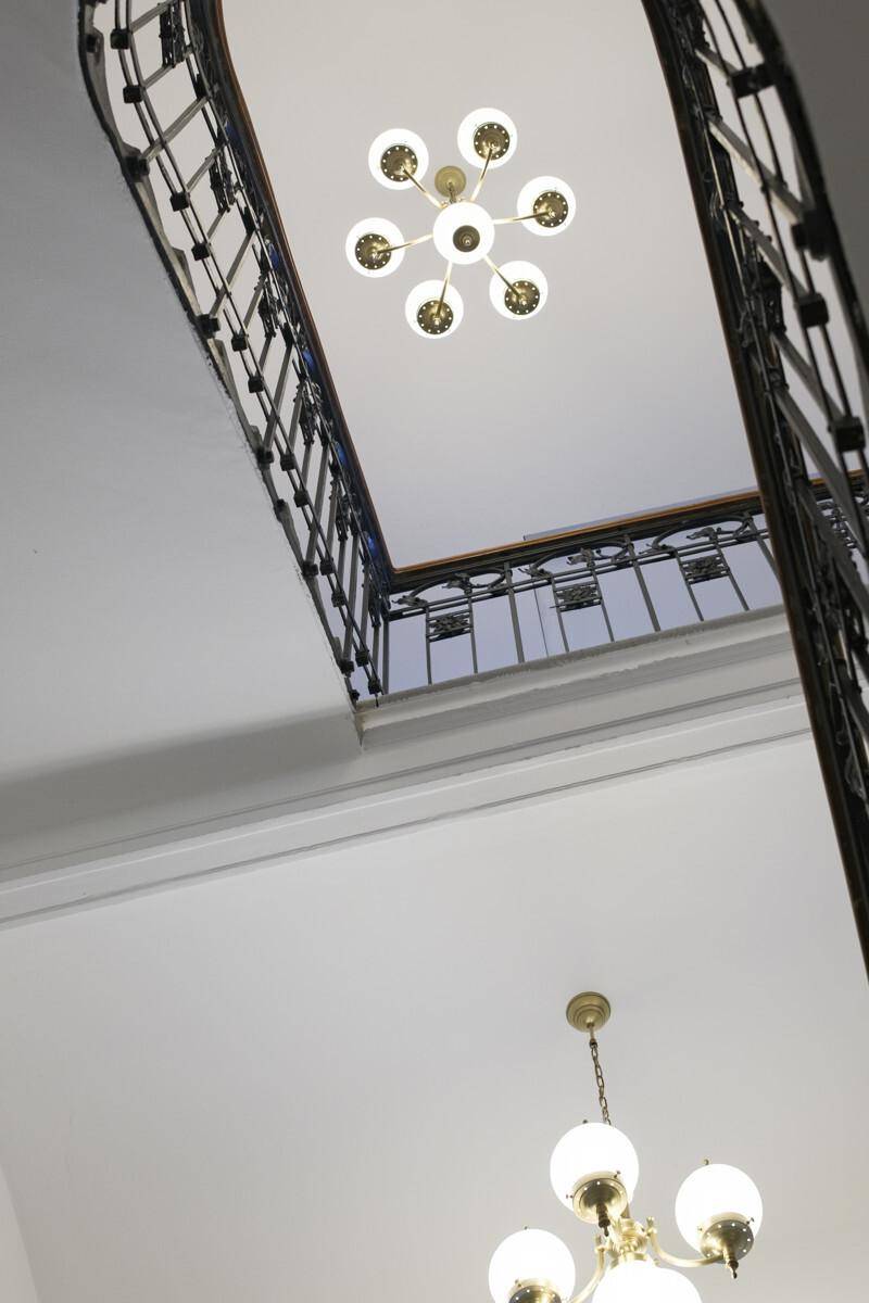 Blick nach oben in ein Treppenhaus mit zwei klassischen Messing-Kronleuchtern und kunstvollem Geländer.