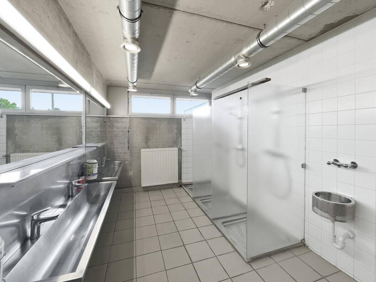 Umkleideraum mit mehreren Duschkabinen, Edelstahlwaschbecken und Fliesenboden, gut beleuchtet.