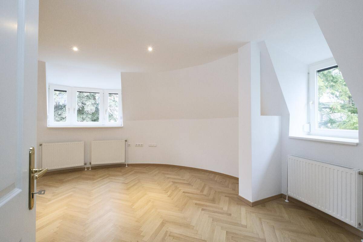 Heller Raum mit Fischgrätparkett, weißen Wänden, mehreren Fenstern und einer geschwungenen Wand.