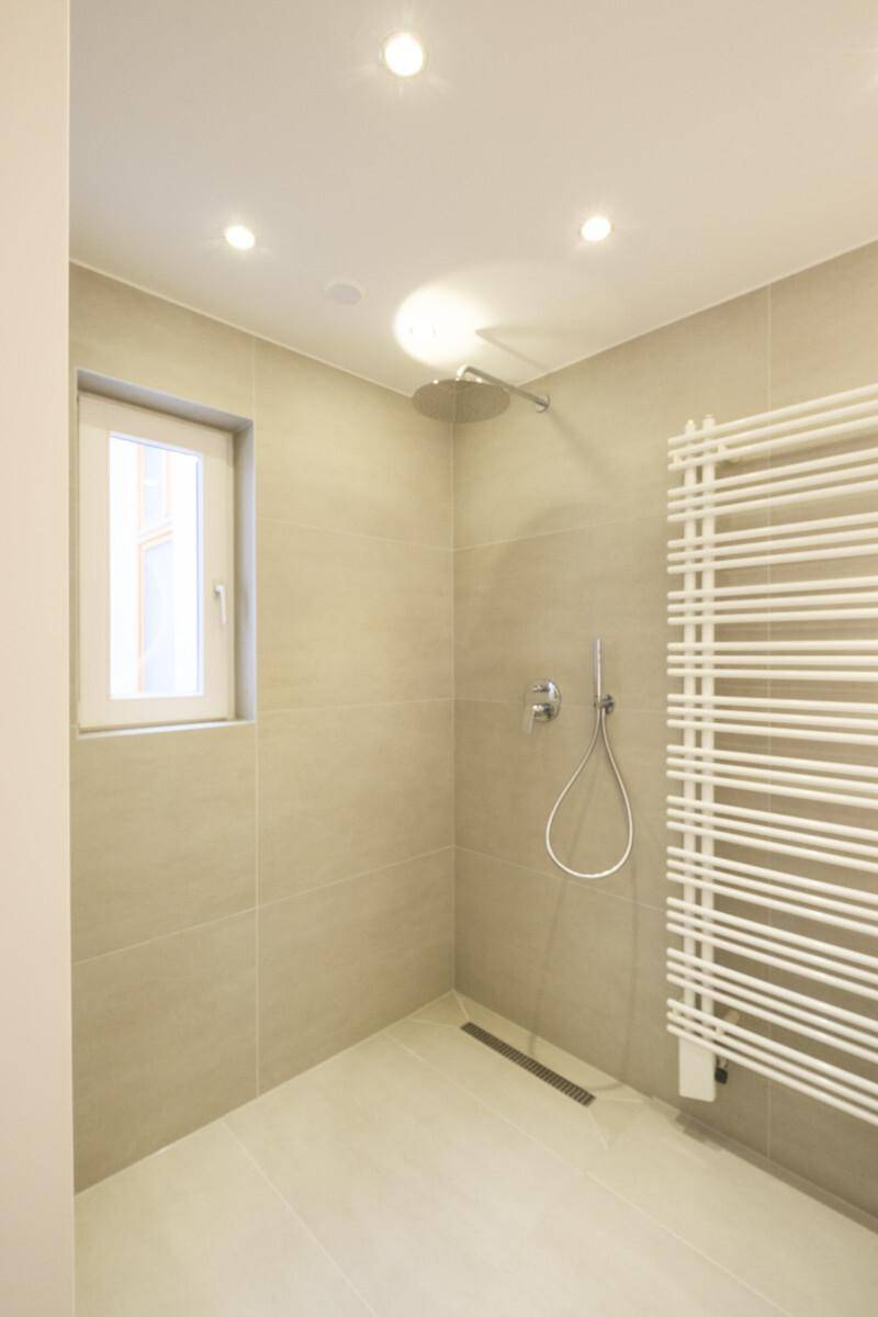 Moderne Duschkabine mit beigen Fliesen, Regendusche, Fenster und praktischem Handtuchheizkörper.