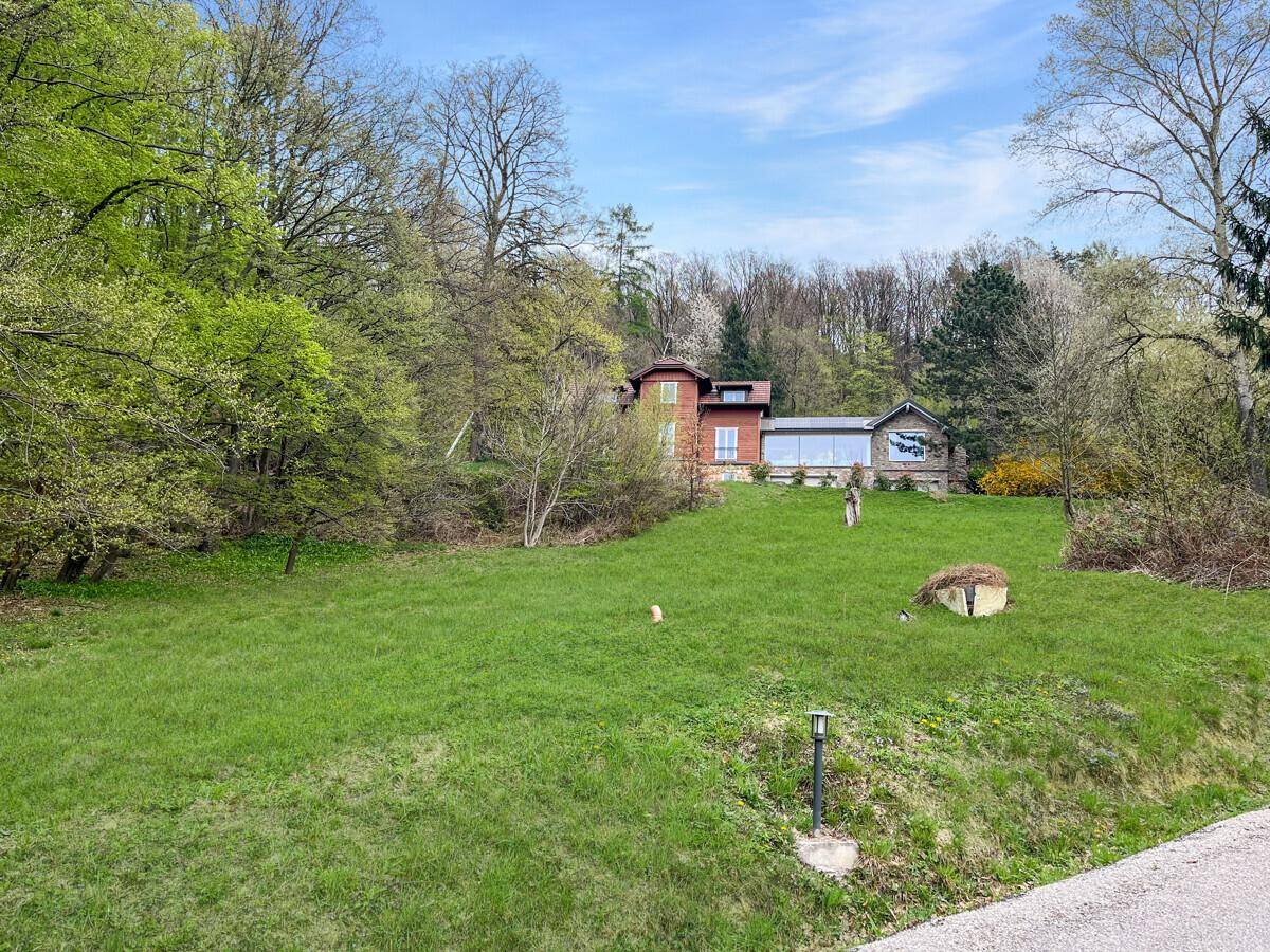 Weitläufige grüne Rasenfläche mit einem Haus im Hintergrund und umgebenden Bäumen.