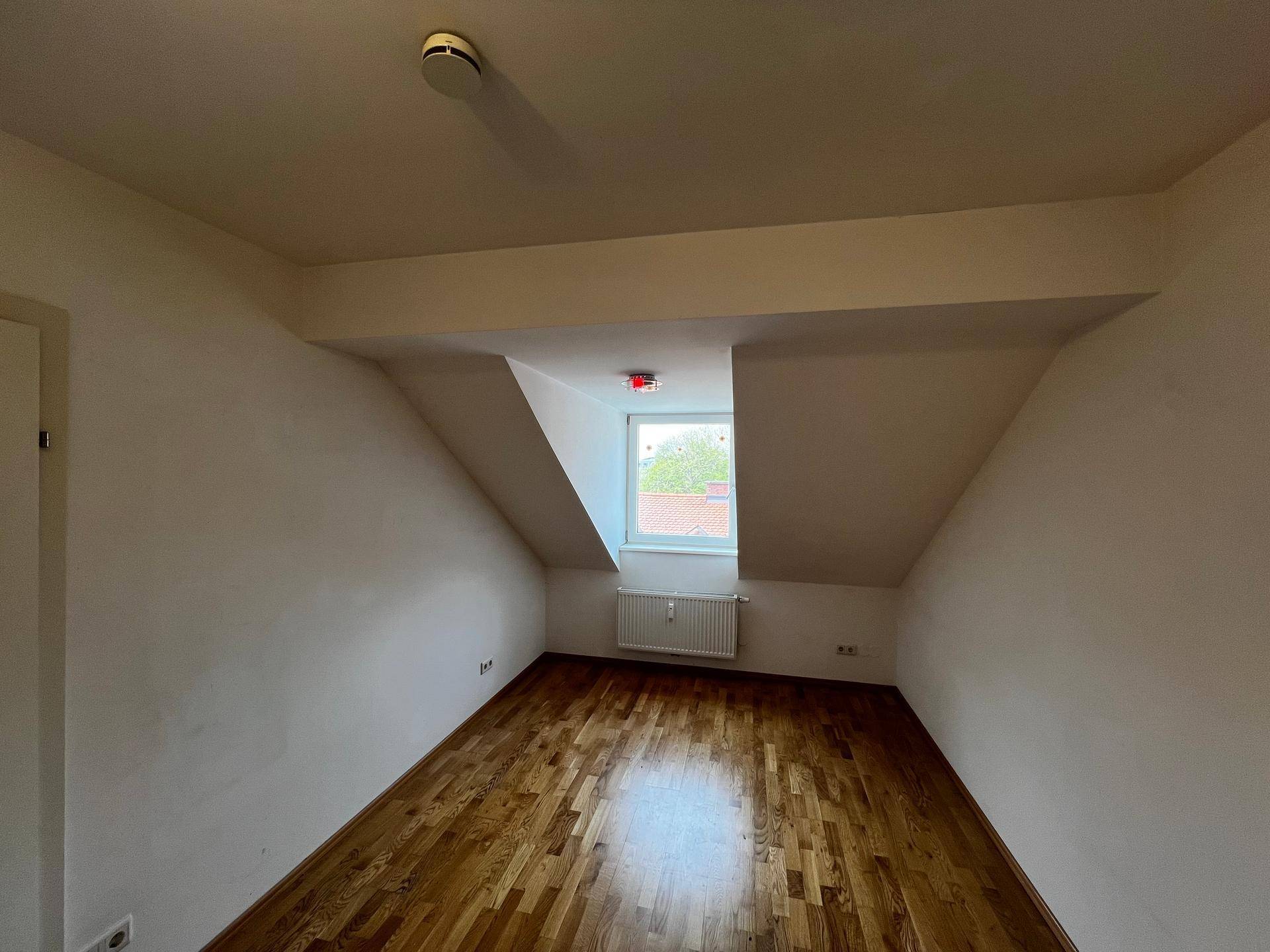 Gemütliches Schlafzimmer mit Holzboden, Dachschrägen und einem Fenster mit Heizkörper.