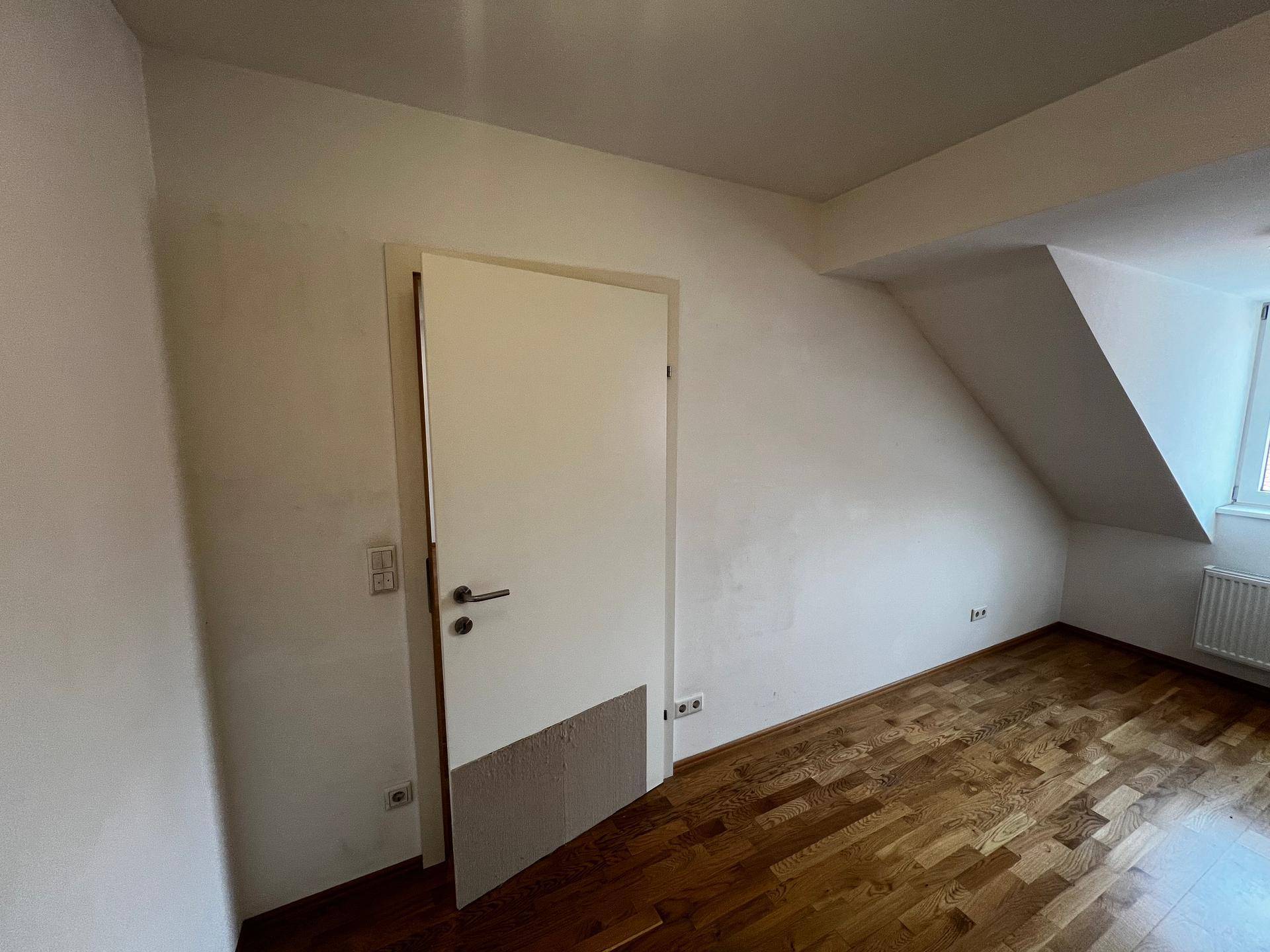 Schlafzimmer mit Holzboden, weiß gestrichenen Wänden und einer Tür, die zu weiteren Räumen führt.