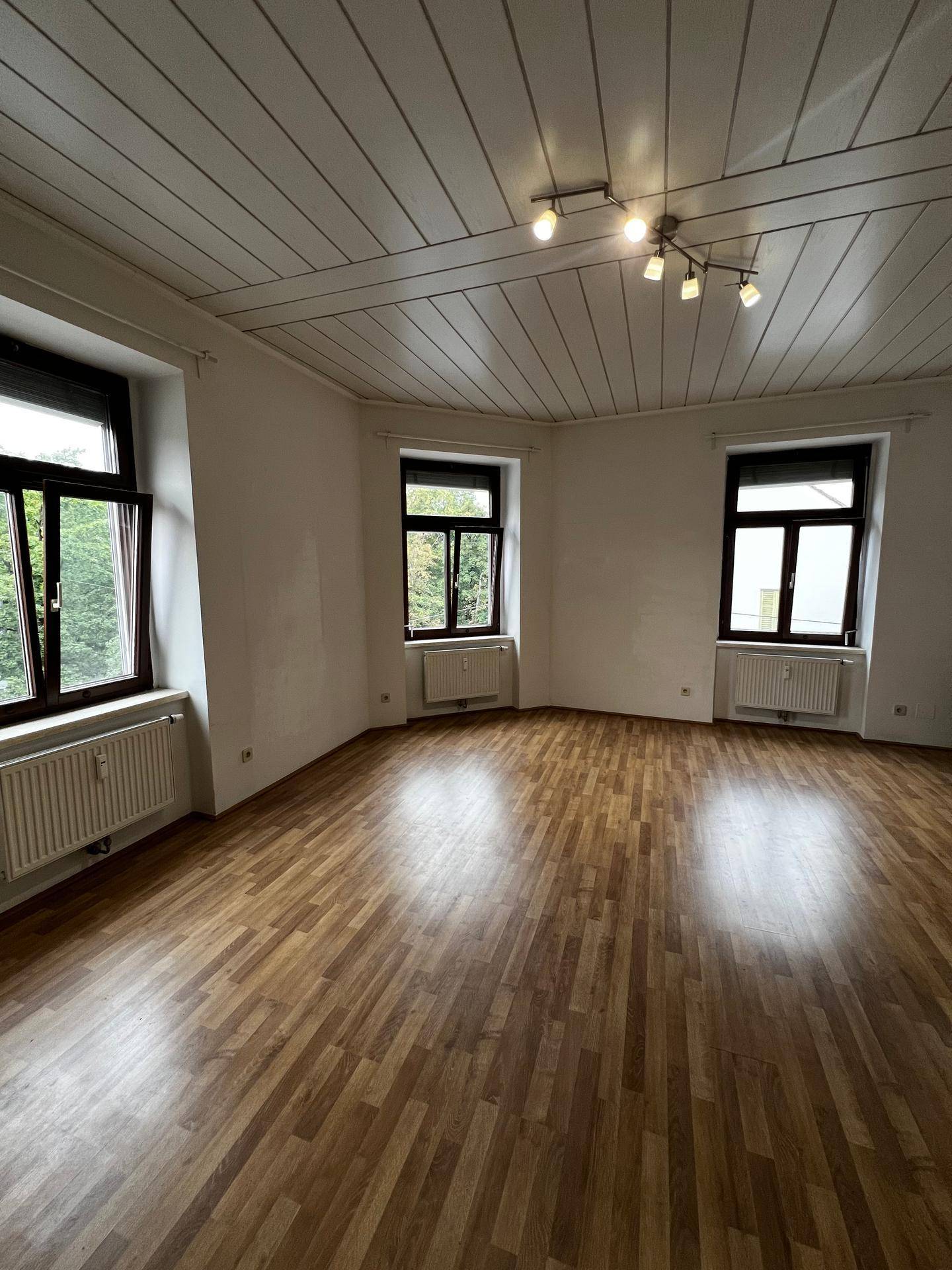 Geräumiger Wohnraum mit Laminatboden, Holzdeckenpaneelen und mehreren Fenstern.