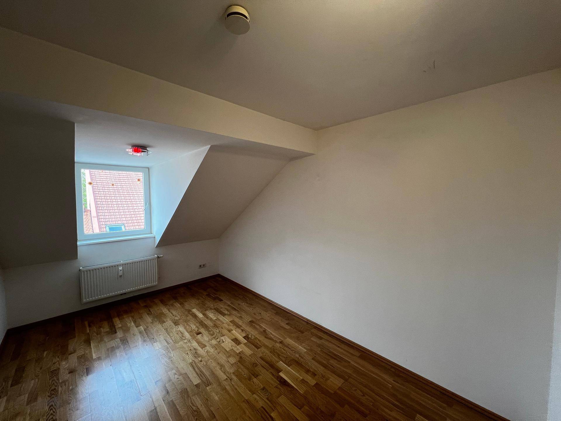 Helles Schlafzimmer mit Holzboden, Dachschrägen und einem Fenster, das Tageslicht hereinlässt.
