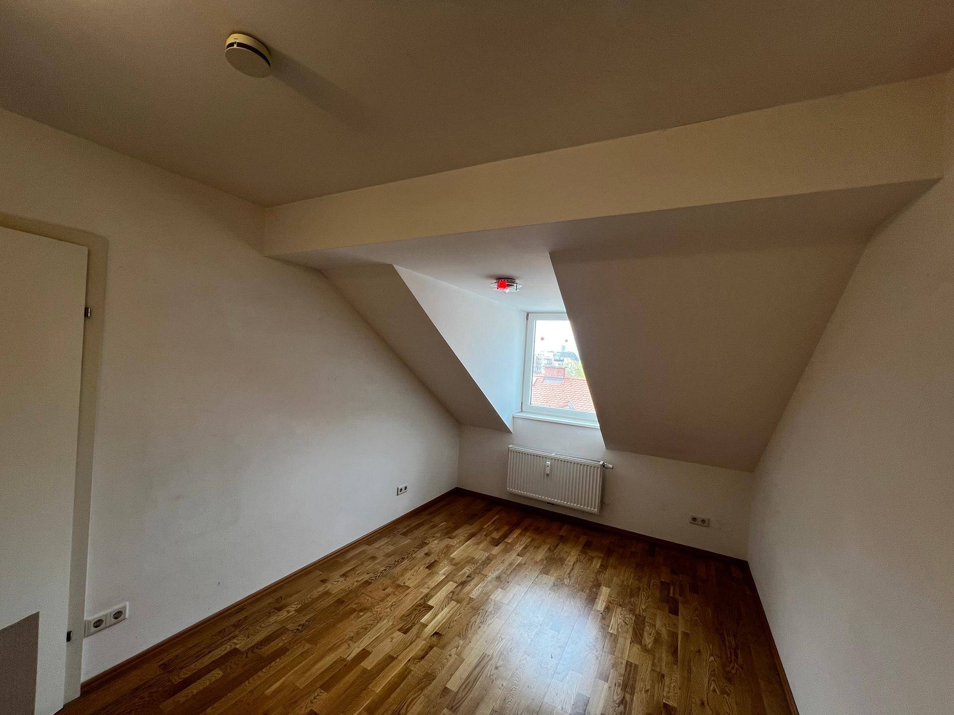Geräumiges Schlafzimmer mit Holzboden, Dachschrägen und einem Fenster mit Heizkörper darunter.