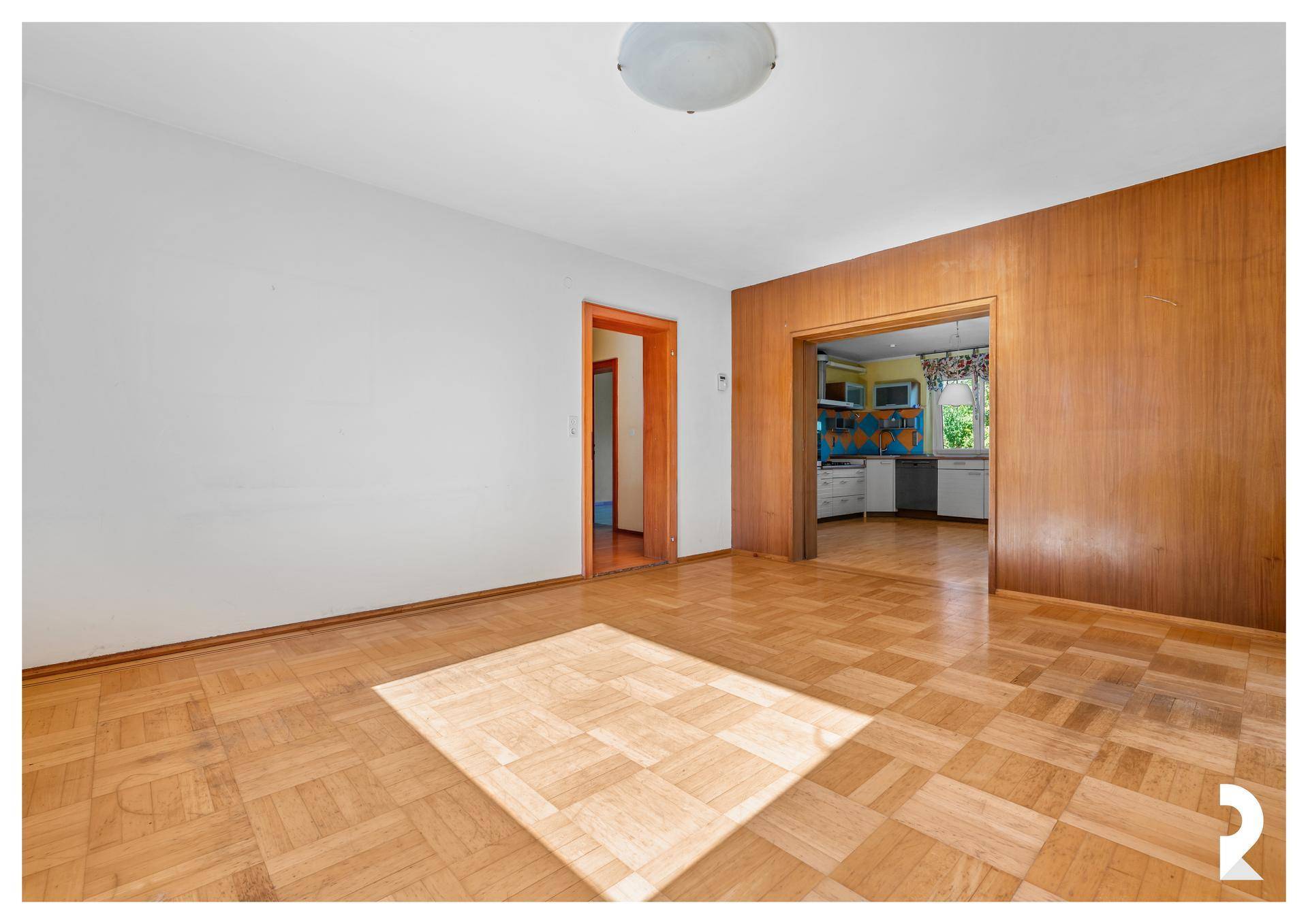 Wohnzimmer mit Parkettboden und Holzvertäfelung, offener Zugang zur Küche im Hintergrund.