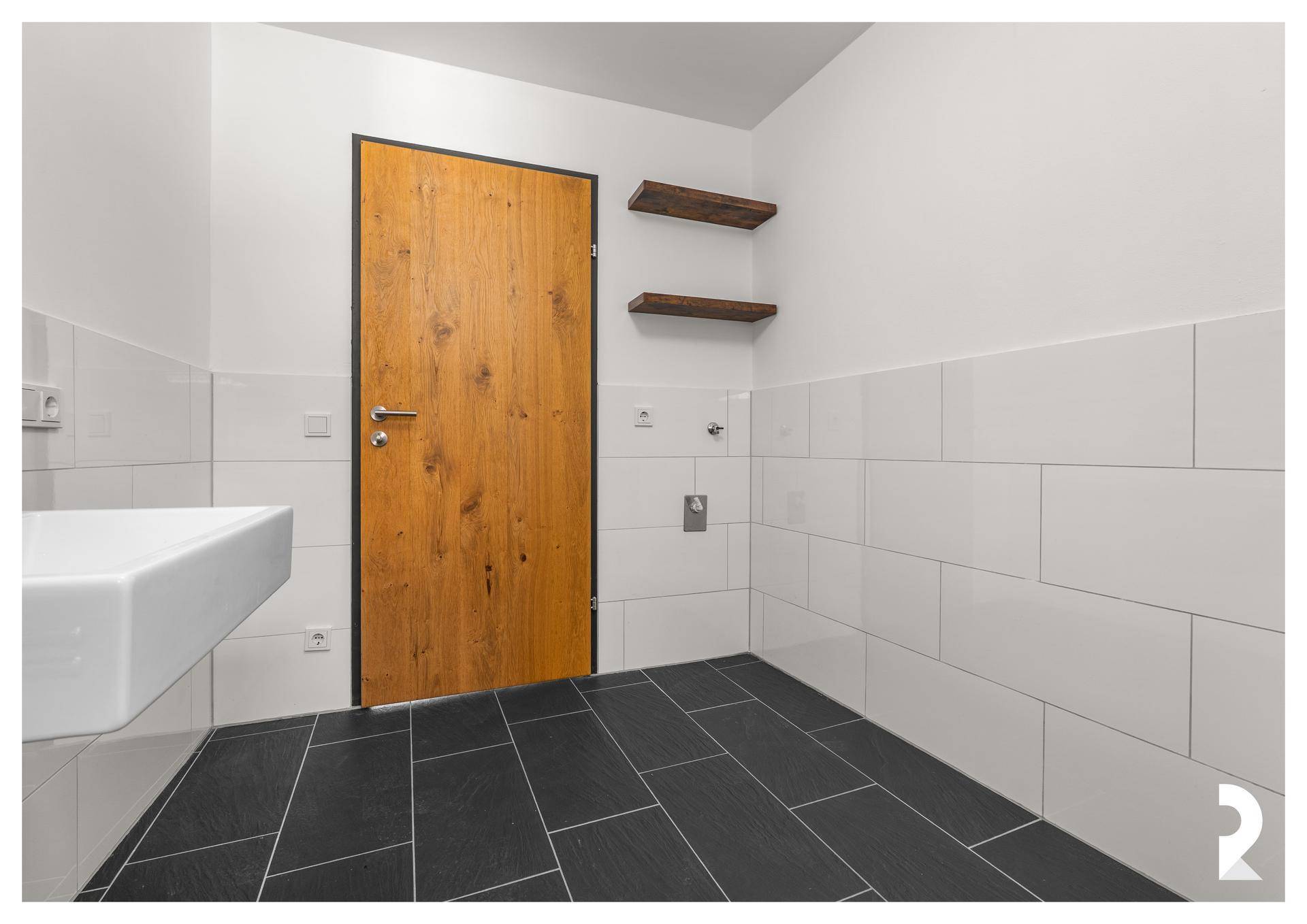 Modernes Badezimmer mit Waschbecken, Holztür, Wandregalen und dunklen Bodenfliesen.