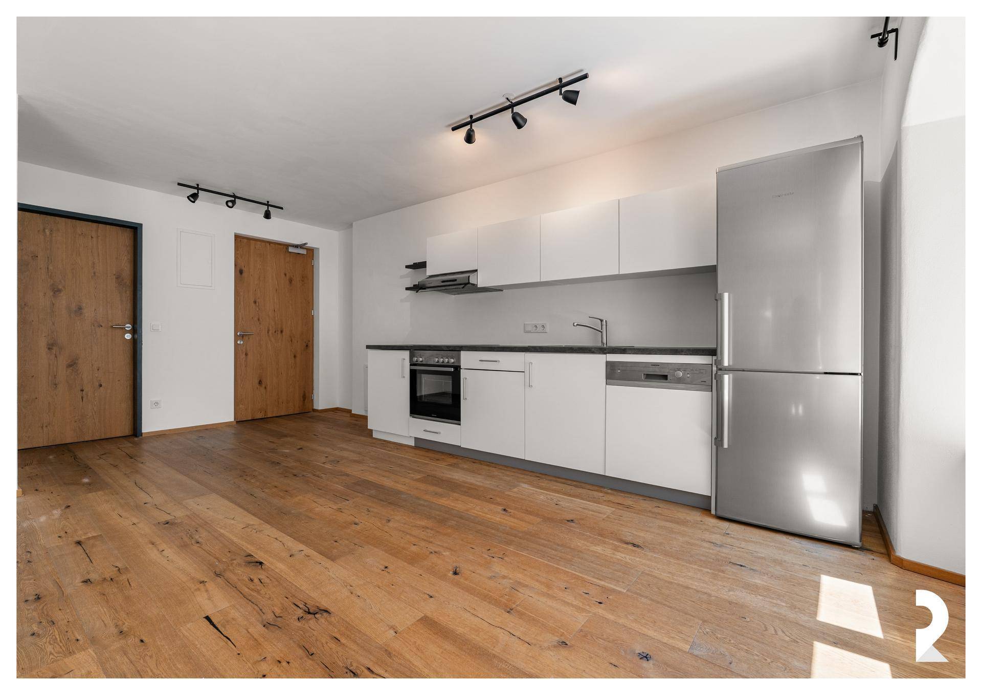 Offener Wohnbereich mit integrierter Küche, Holzboden und zwei Holztüren, die zu weiteren Räumen führen.